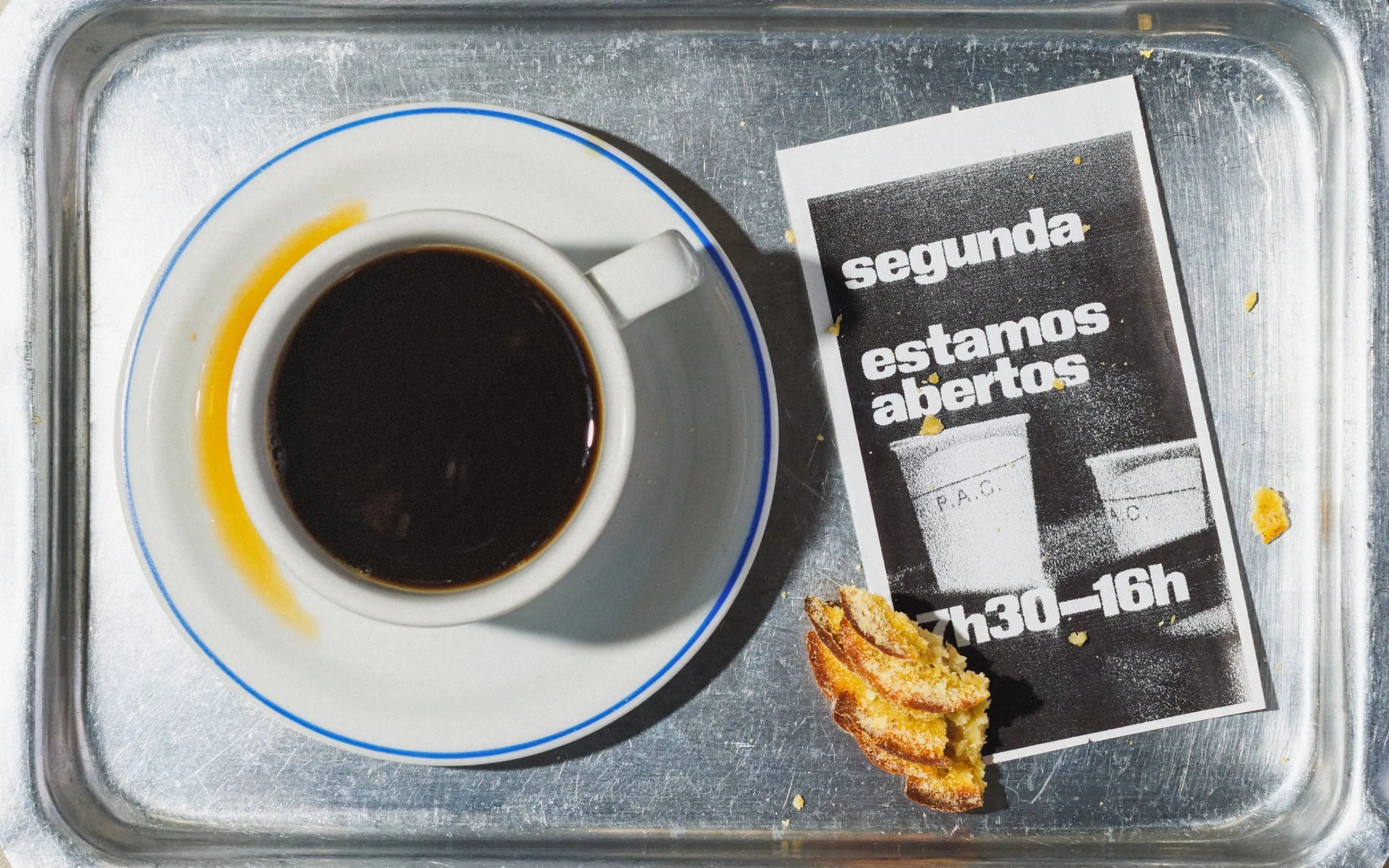 Bandeja de metal con un café solo, un trozo de bollo y un folleto en blanco y negro que dice «segunda, estamos abiertos»