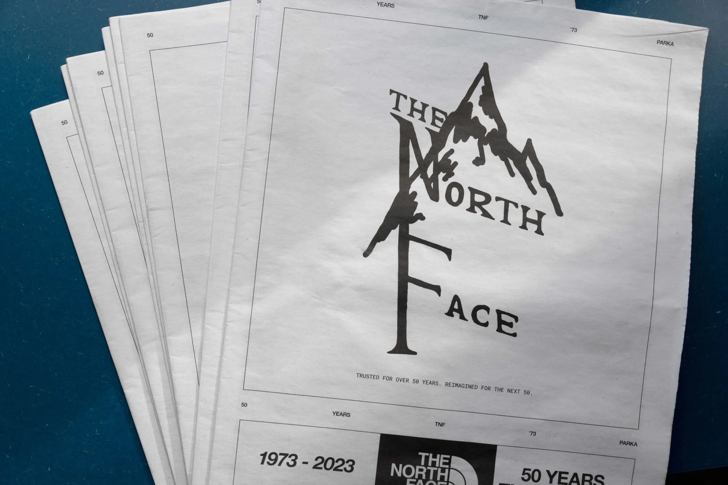Varios ejemplares de un fanzine en papel de periódico sobre una mesa azul; en la portada, el logotipo antiguo de The North Face dibujado a mano con una montaña y el texto «1973–2023, 50 years»