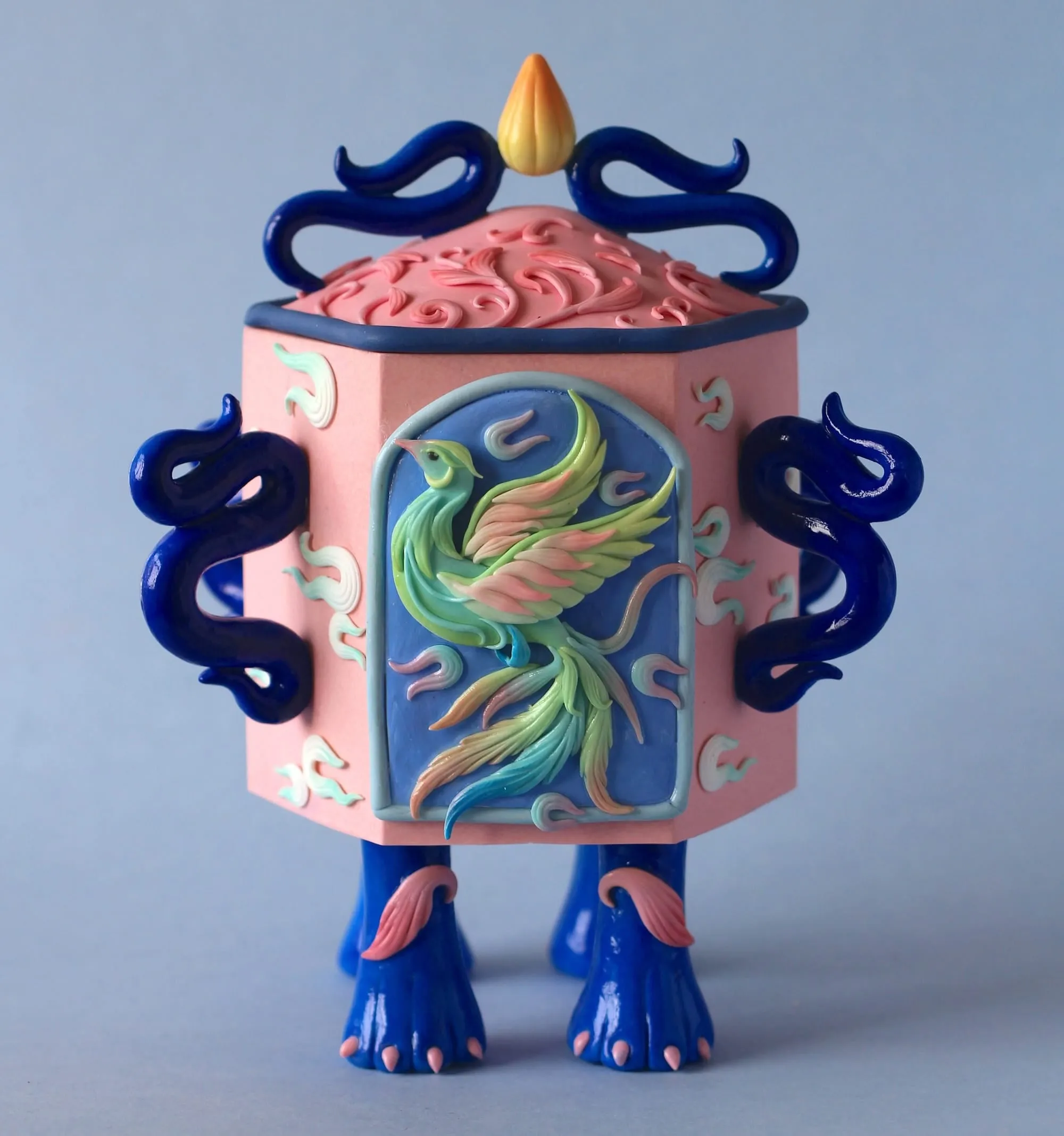 Botella hexagonal rosa con un fénix verde y rosa en relieve sobre un panel azul, asas azules retorcidas y patas azules con uñas rosas
