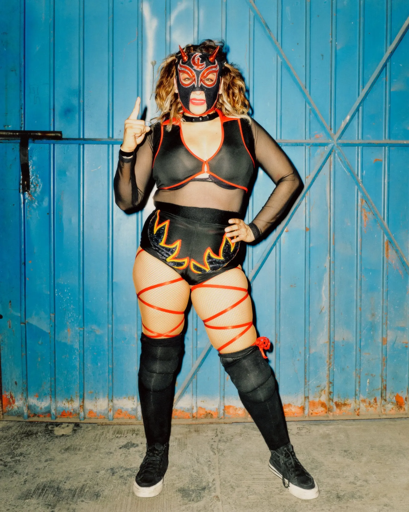 Luchadora con máscara negra y roja de cuernos posa señalando hacia arriba frente a un portón metálico azul