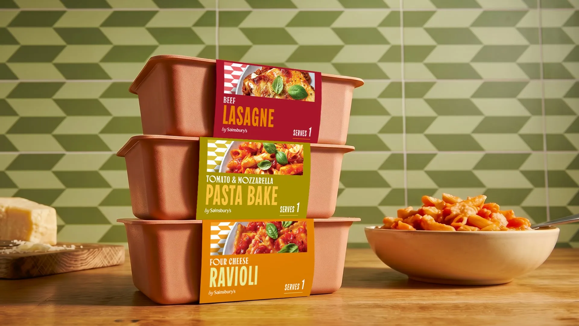 Tres bandejas color terracota apiladas con bandas de papel de lasaña, pasta al horno y ravioli, ante una pared de azulejos verdes en zigzag