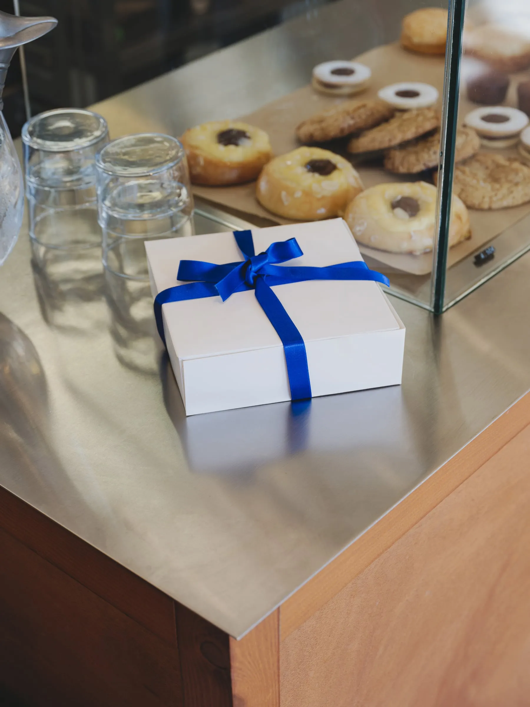 Caja blanca de pastelería cerrada con un lazo azul cobalto, sobre una barra de acero junto a una vitrina con bollos