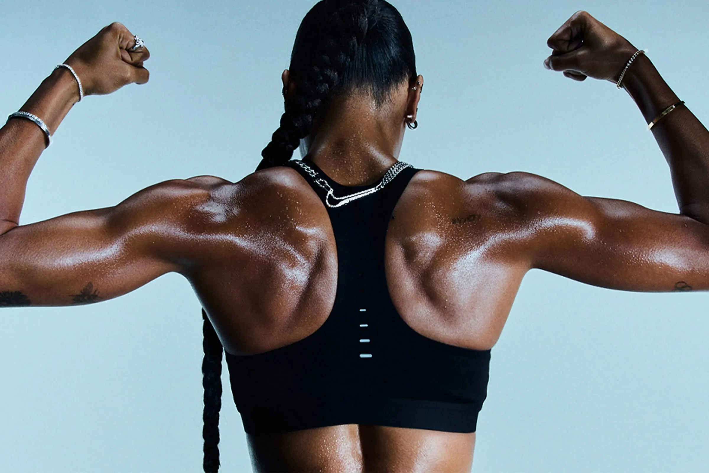 Mujer de espaldas con los brazos flexionados, con la cadena de la medalla al cuello y una pulsera en cada muñeca