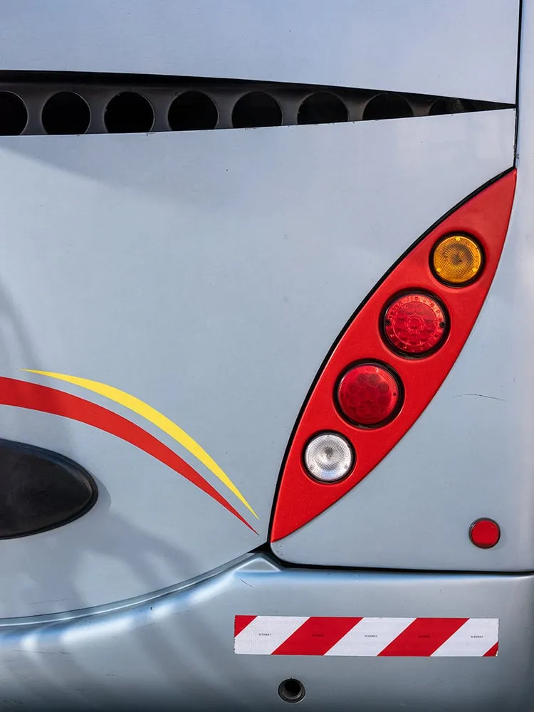 Trasera plateada de un autobús con una franja roja y amarilla, un piloto rojo en forma de lágrima y una banda de rayas rojas y blancas