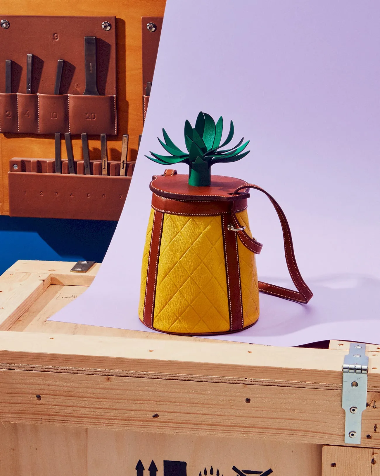 Bolso de piel de Hermès en forma de piña, amarillo acolchado con asas marrones y hojas de cuero verde, sobre un cajón de madera y un fondo lila, con herramientas de marroquinería colgadas detrás
