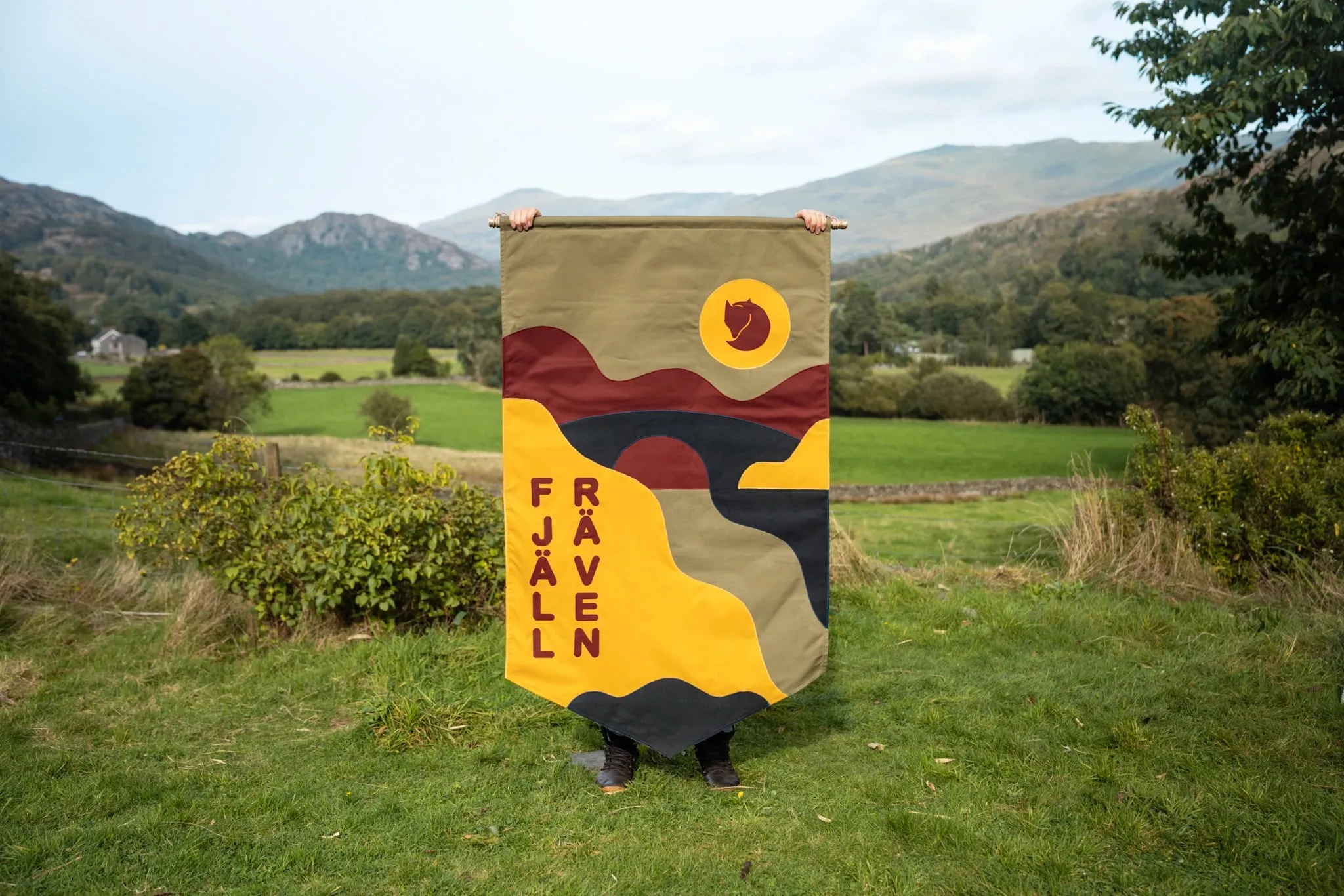 Bandera vertical de tela con un valle, un río y el zorro de Fjällräven sobre un sol amarillo, con el nombre de la marca en columnas