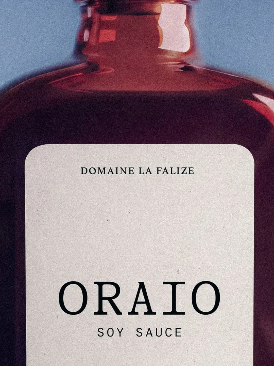 Primer plano de la botella ámbar de Oraio con etiqueta de papel crema: arriba «Domaine La Falize», abajo «Oraio» en mayúsculas con remates y «Soy Sauce»