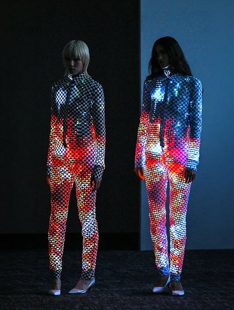 Dos modelos de cuerpo entero con trajes de cuadros y escamas cubiertos de luces de colores, sobre una moqueta oscura y zapatos de tacón claros