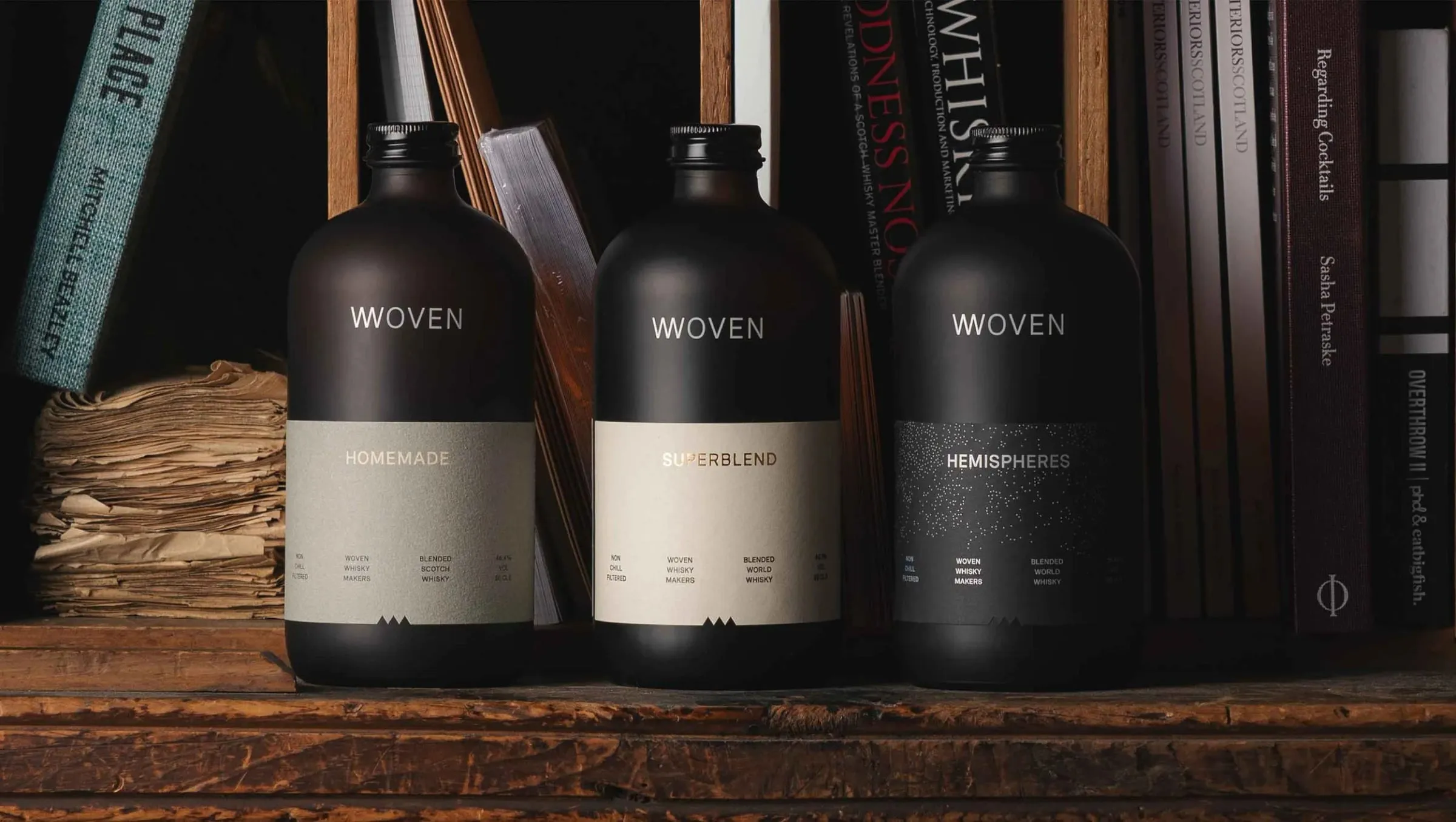 Tres botellas negras de Woven en una estantería de madera con libros, con etiquetas gris verdoso, crema y negra con puntos