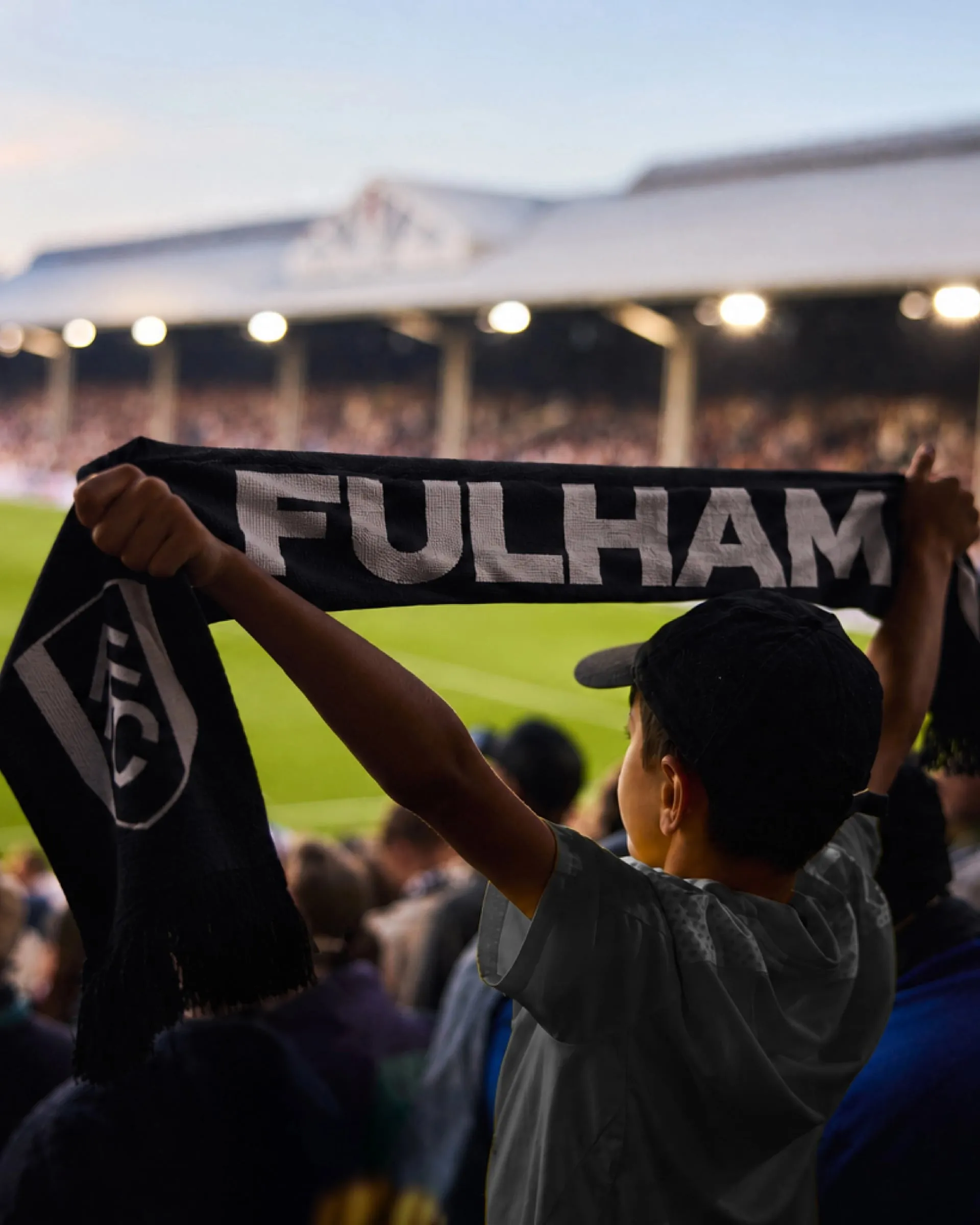 Un niño levanta una bufanda negra con FULHAM en letras blancas y el escudo del club, con la grada de Craven Cottage desenfocada al fondo