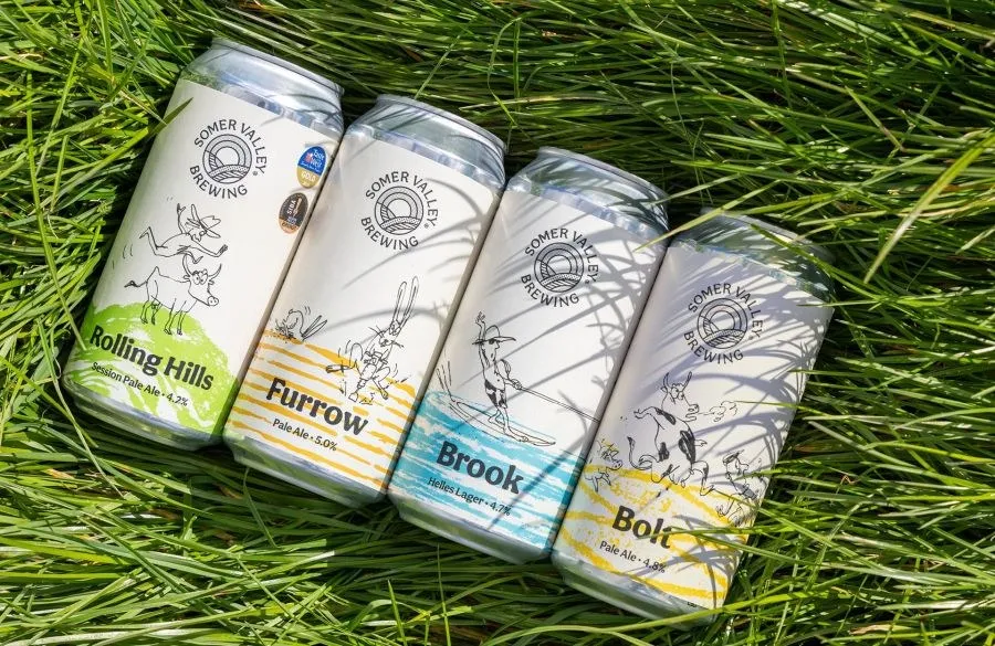 Cuatro latas de cerveza blancas de Somer Valley Brewing con dibujos de animales, tumbadas sobre hierba verde