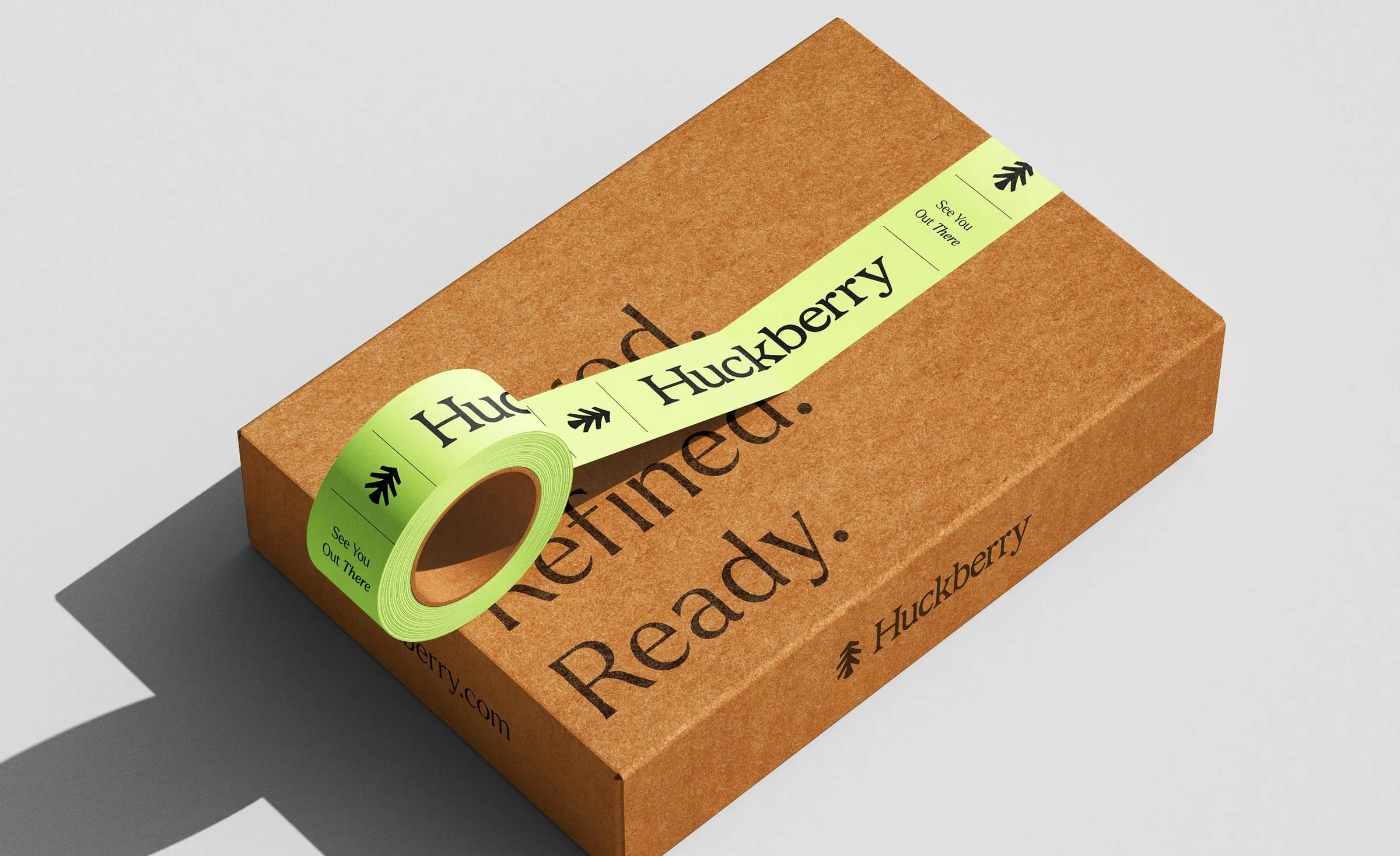Caja de cartón kraft con el lema «Rugged. Refined. Ready.» impreso y un rollo de cinta adhesiva verde lima con el logotipo de Huckberry y el texto «See You Out There»