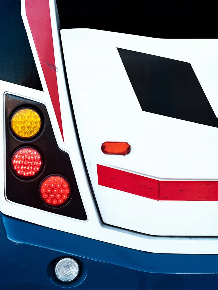Trasera blanca y azul de un autobús con una ventana negra en forma de rombo, una franja roja y pilotos naranjas y rojos
