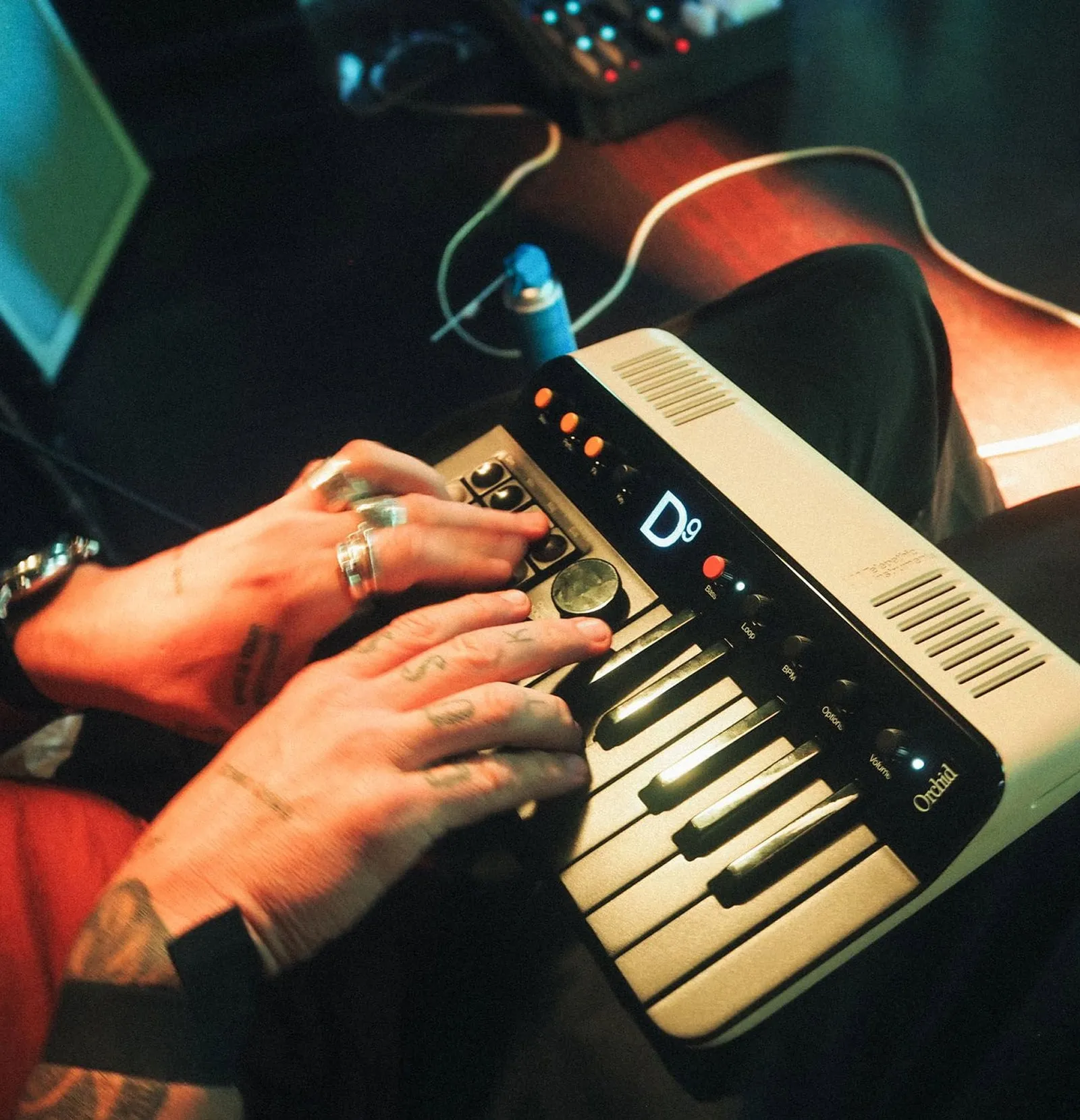 Dos manos con anillos tocan el teclado de Orchid; la pantalla negra muestra un «D9» en letras blancas y alrededor se ven mandos rotulados como «Loop» o «BPM»