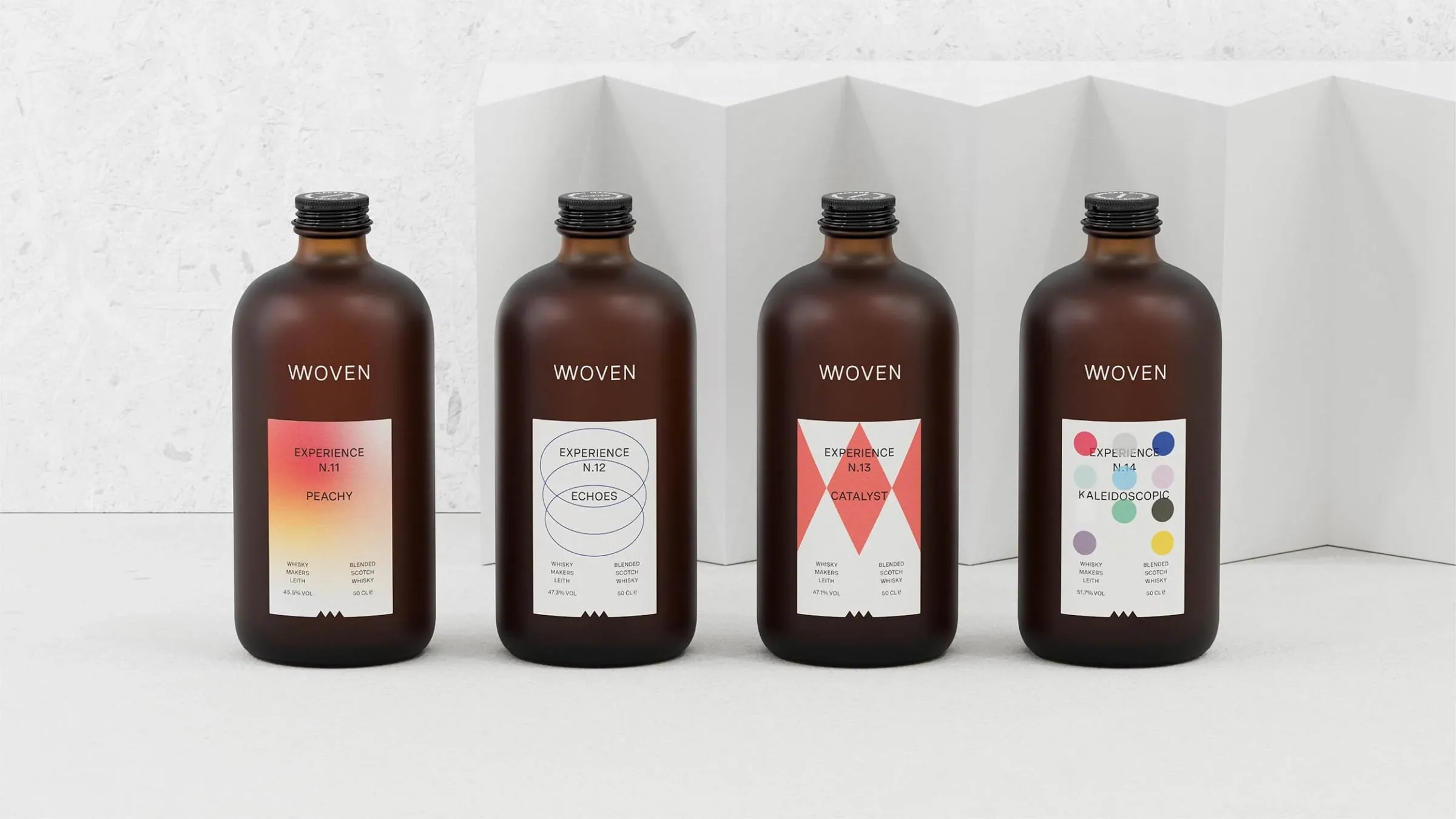 Cuatro botellas marrones esmeriladas de whisky Woven en fila, cada una con una etiqueta de color distinto: degradado rojo, círculos azules, rombos rojos y puntos de colores