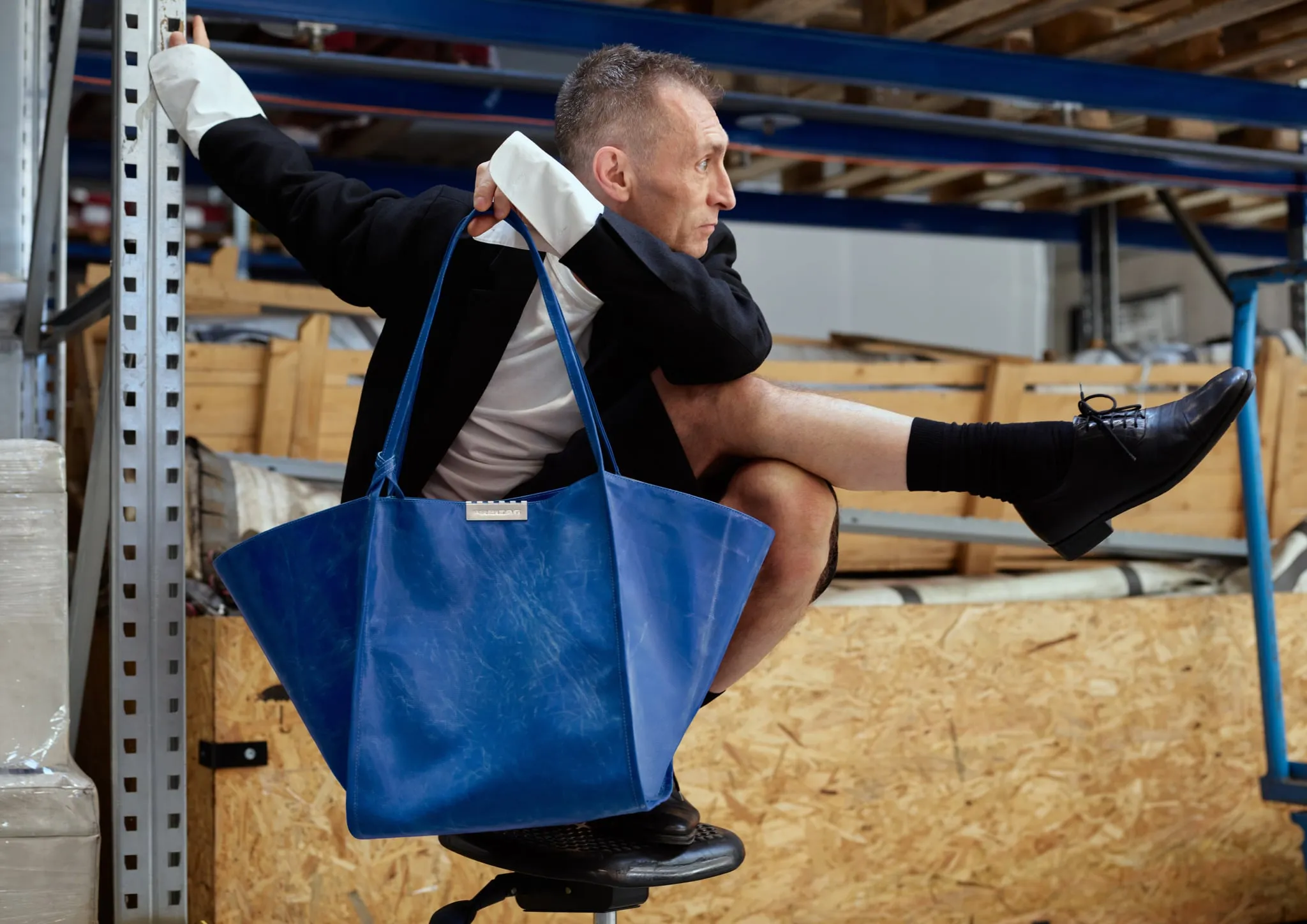 Persona agachada en un almacén con un tote azul grande colgado del brazo, entre estanterías metálicas y cajas de madera