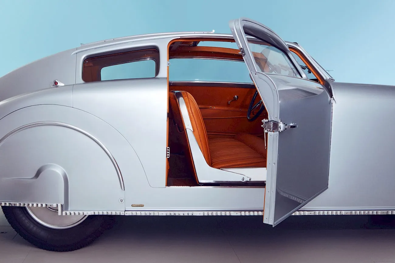 Lateral de un coche antiguo plateado con la puerta abierta y el interior tapizado en cuero color coñac, sobre un fondo azul claro