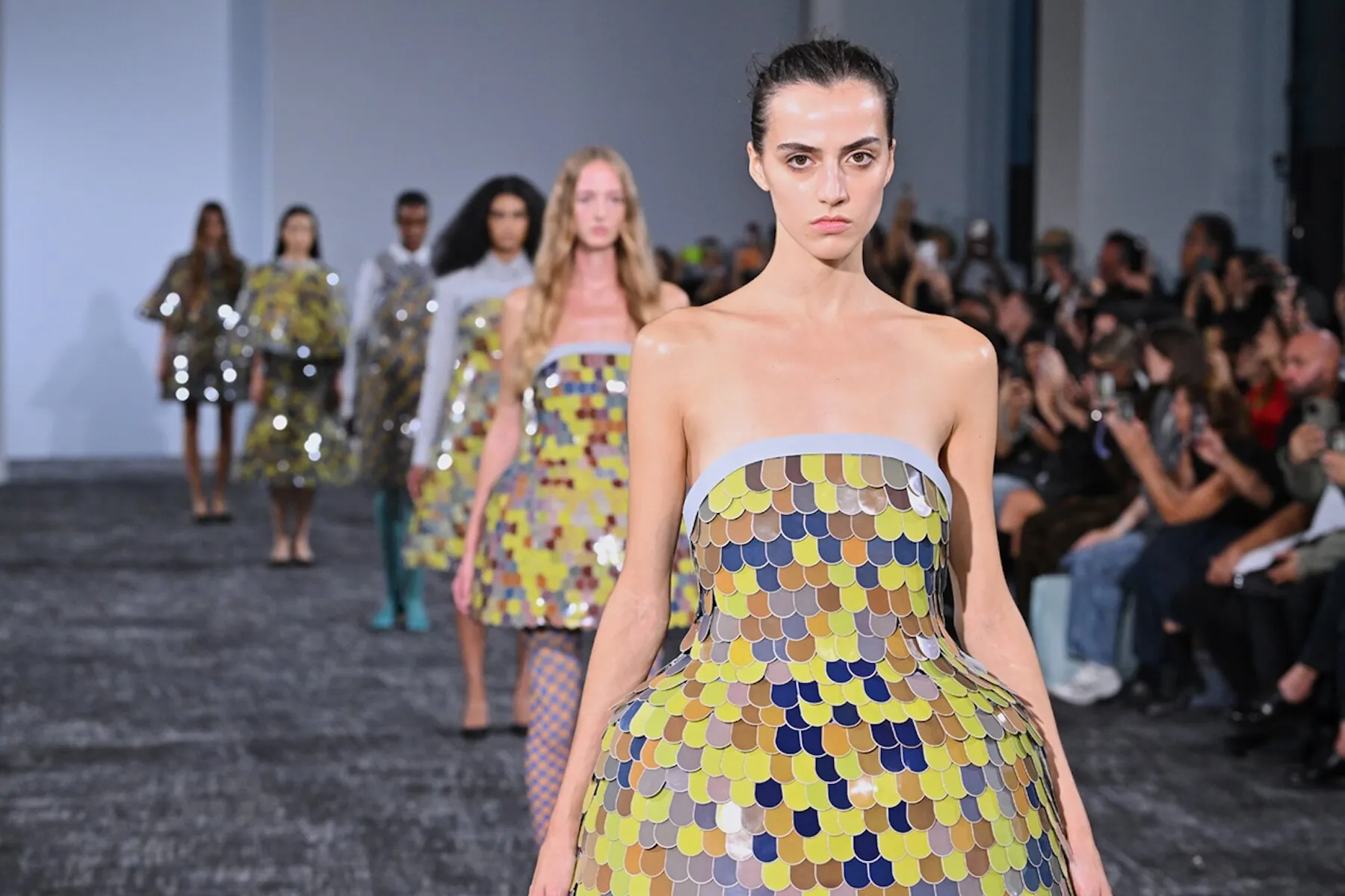 Modelo de frente con un vestido sin tirantes cubierto de escamas amarillas, azules, ocres y grises, y otras modelos detrás en la pasarela