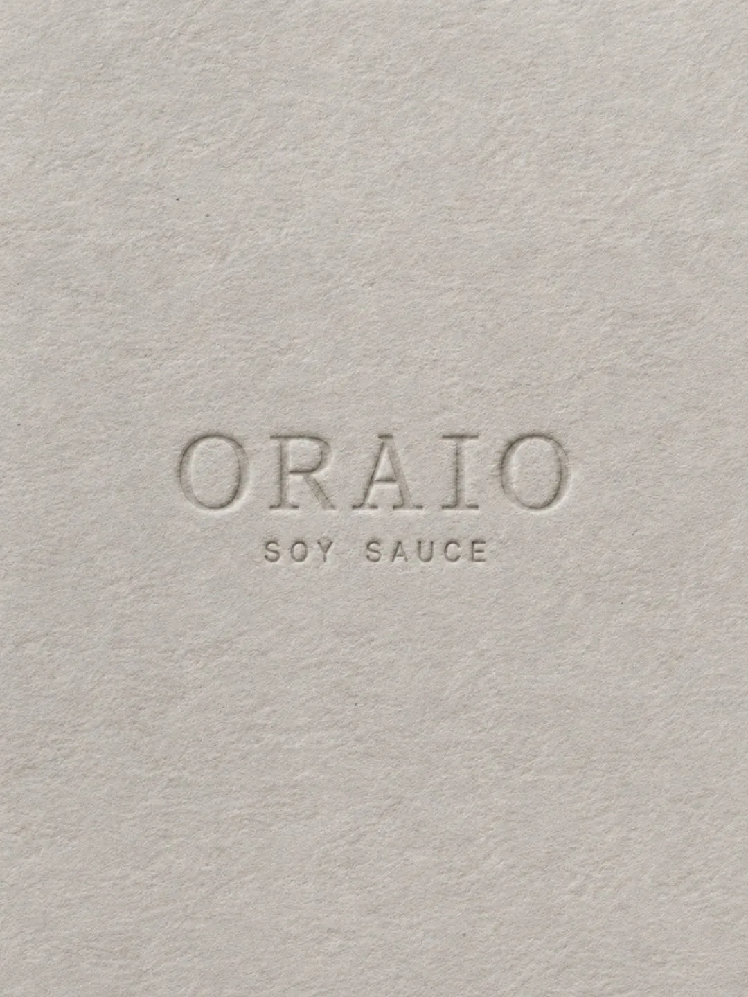 Logotipo de Oraio estampado en seco, sin tinta, sobre papel gris con textura