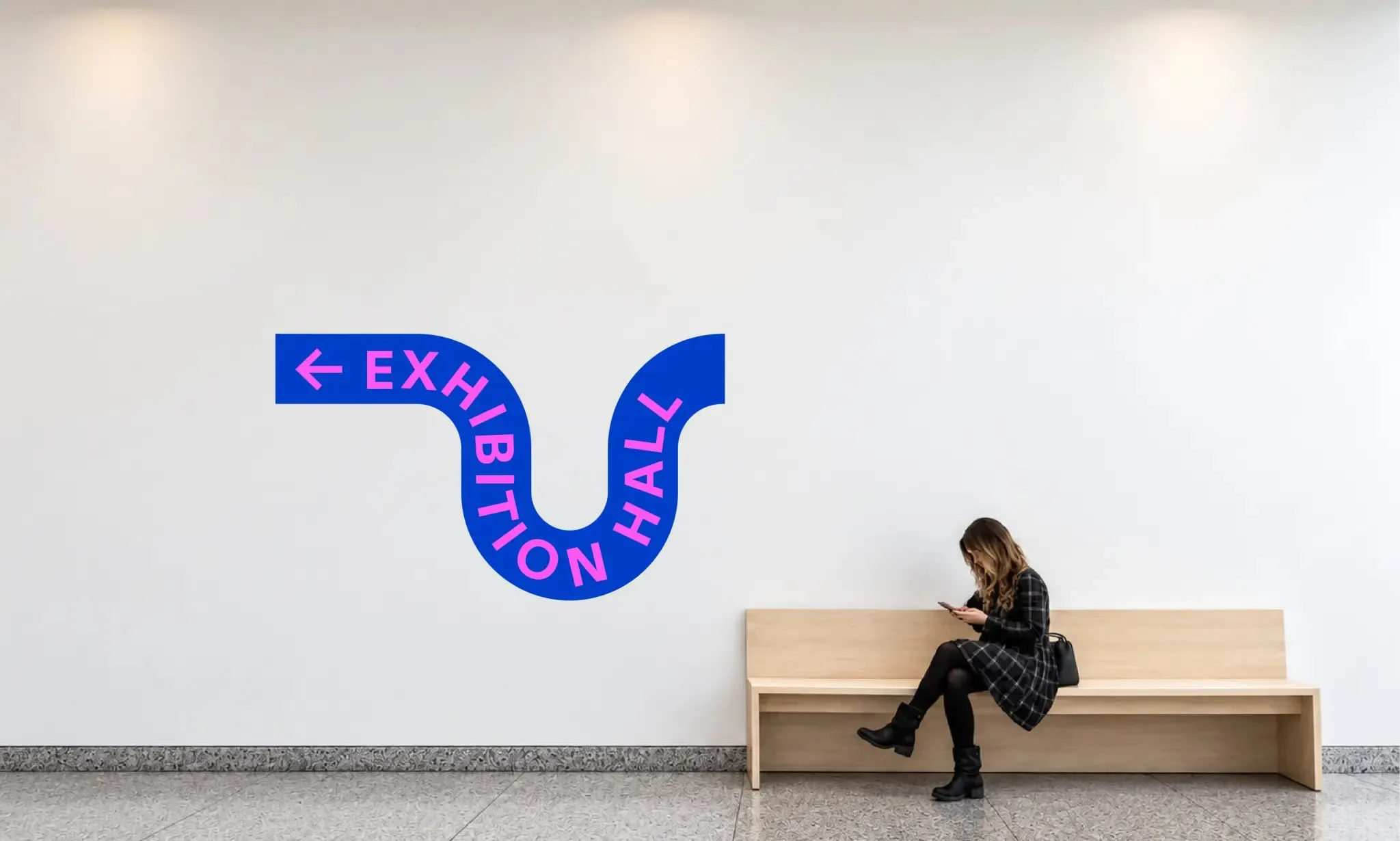 Rótulo azul de una pared blanca con una flecha y las letras «Exhibition Hall» siguiendo una curva en forma de S, junto a una mujer sentada en un banco