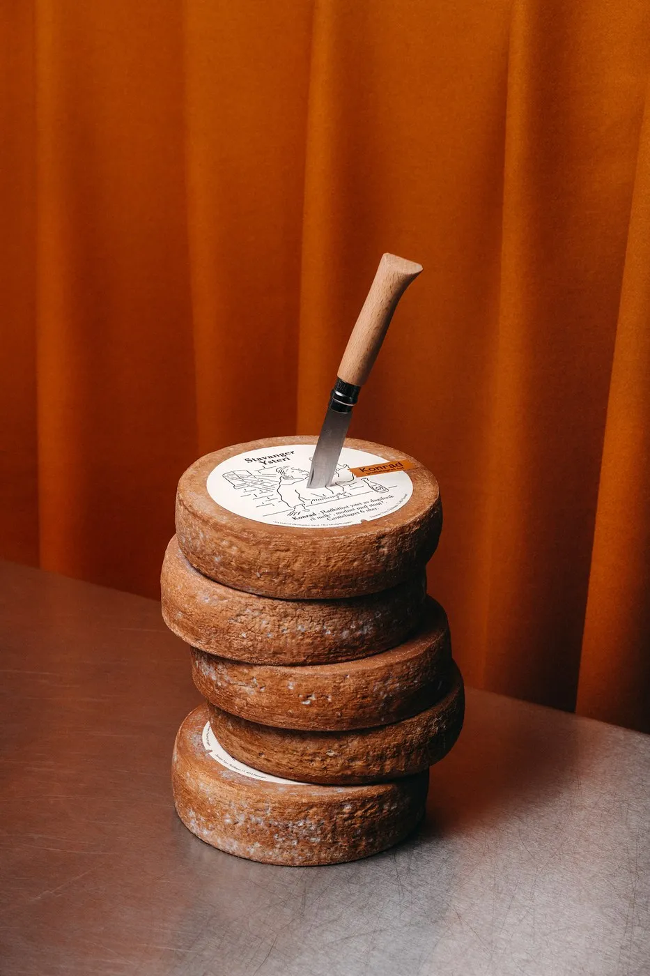 Pila de ruedas de queso oscuras con un cuchillo de mango de madera clavado en la de arriba, sobre un telón naranja