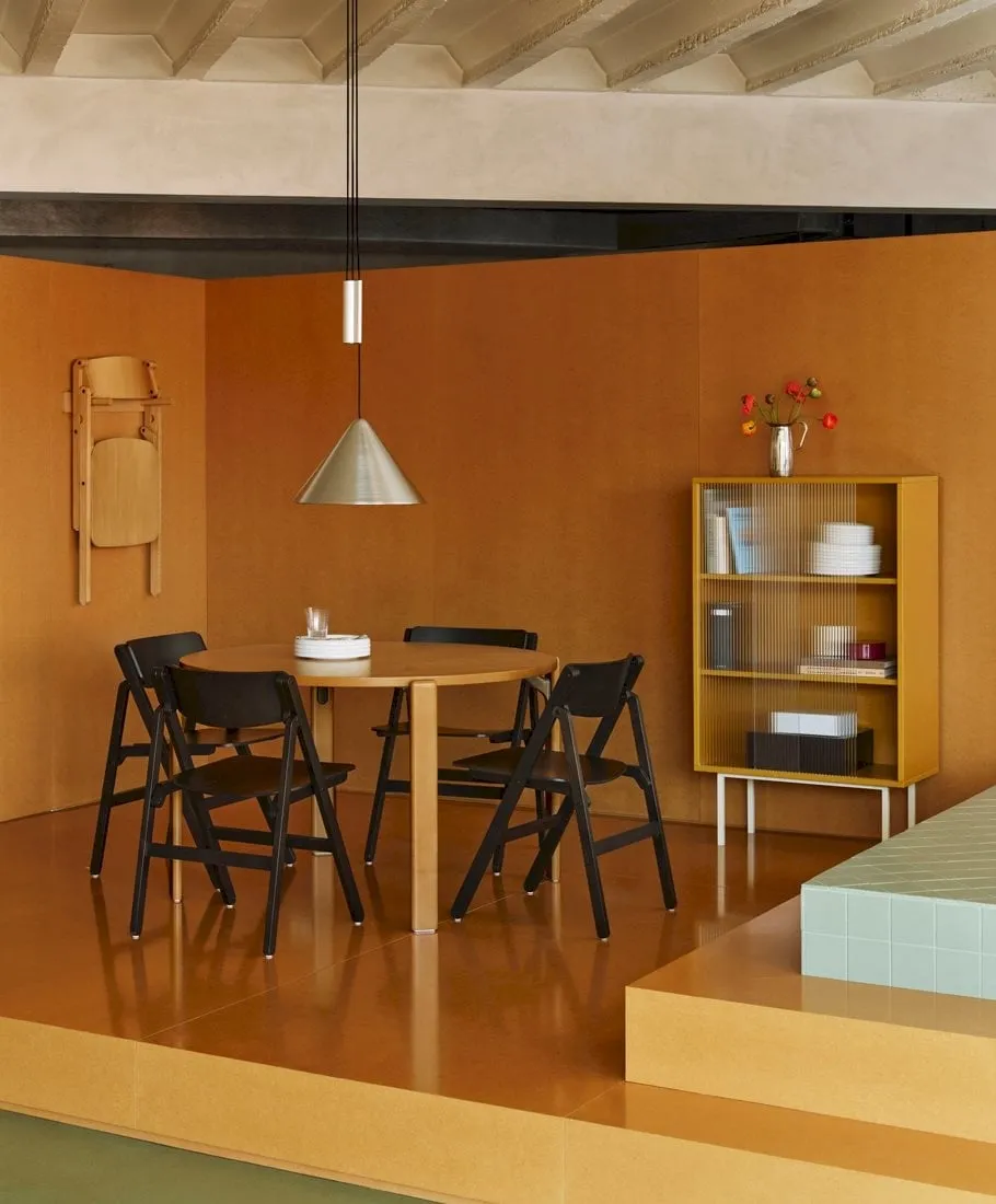 Comedor en tonos ocres con una mesa redonda rodeada de sillas Backflip negras y una silla plegada colgada de la pared junto a una lámpara colgante