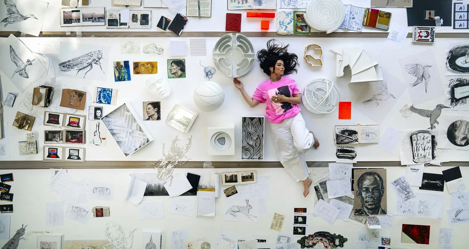 Es Devlin tumbada sobre una mesa blanca rodeada de maquetas, dibujos y cuadernos vistos desde arriba