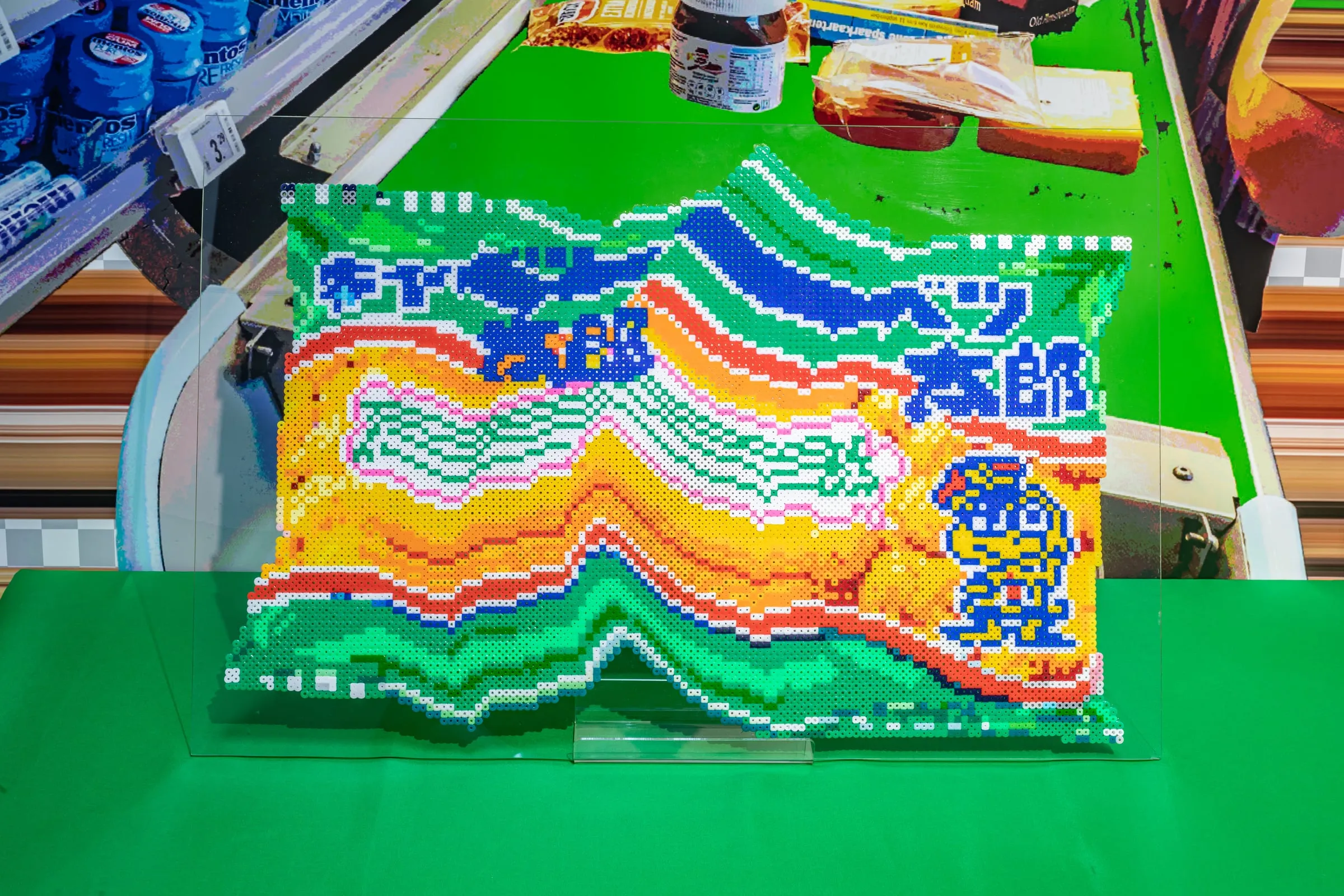 Bolsa de snacks hecha de cuentas de colores sobre una mesa verde, con ondas de naranja, azul y verde y un personaje de dibujos en una esquina