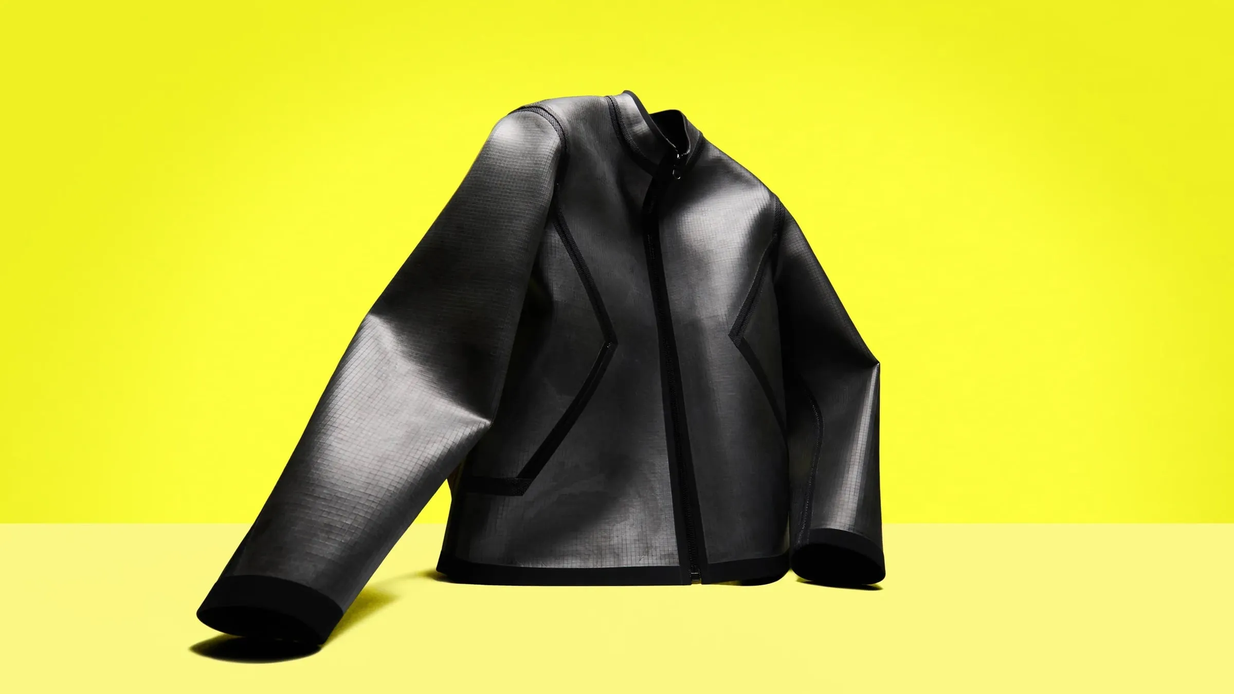 Chaqueta negra corta de pizarra con cremallera central y costuras selladas con cinta, sobre fondo amarillo
