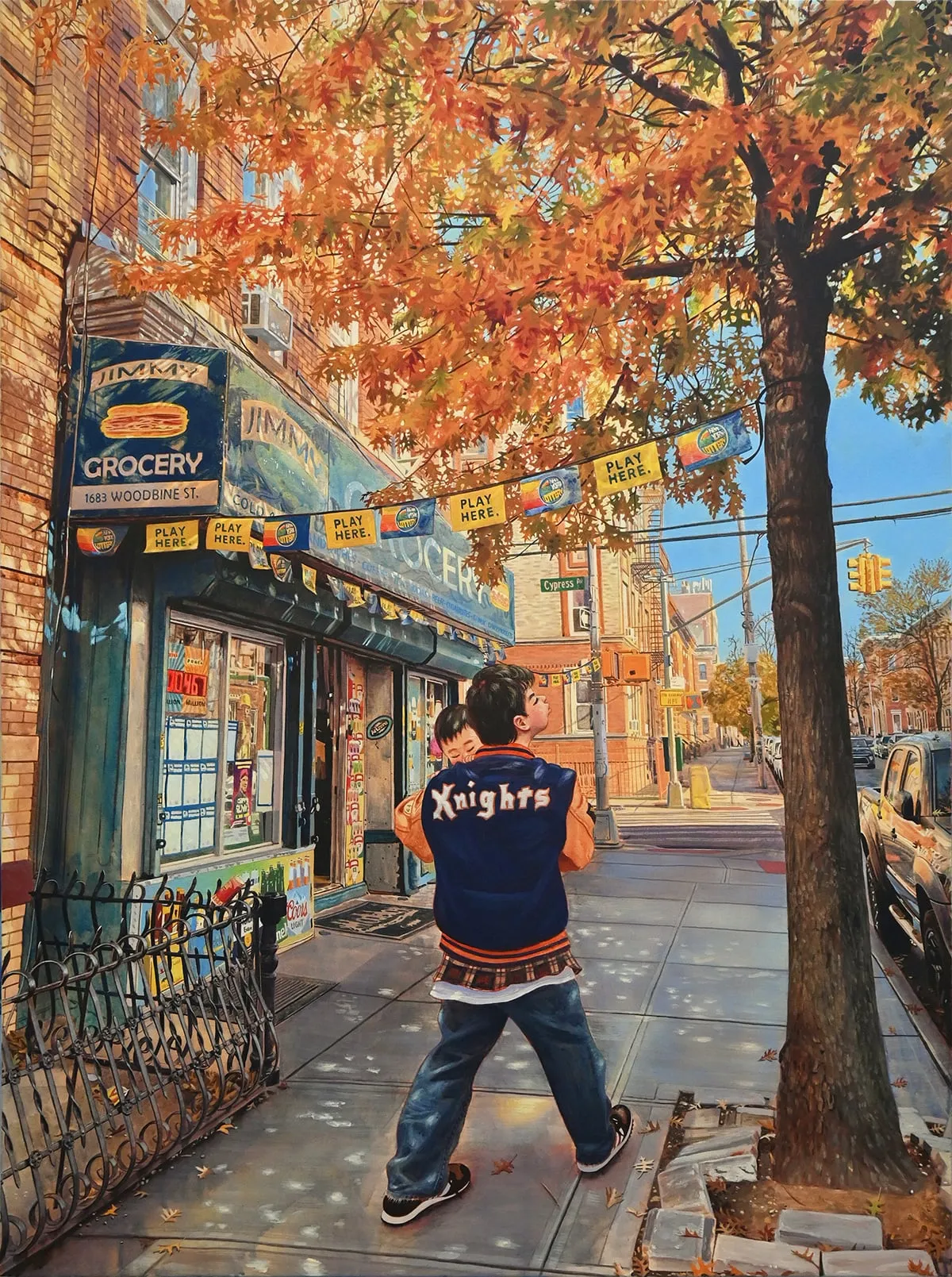 Calle de Brooklyn en otoño: un chico con una chaqueta azul que pone «Knights» lleva a un niño en brazos bajo un árbol de hojas naranjas, junto a una tienda de comestibles