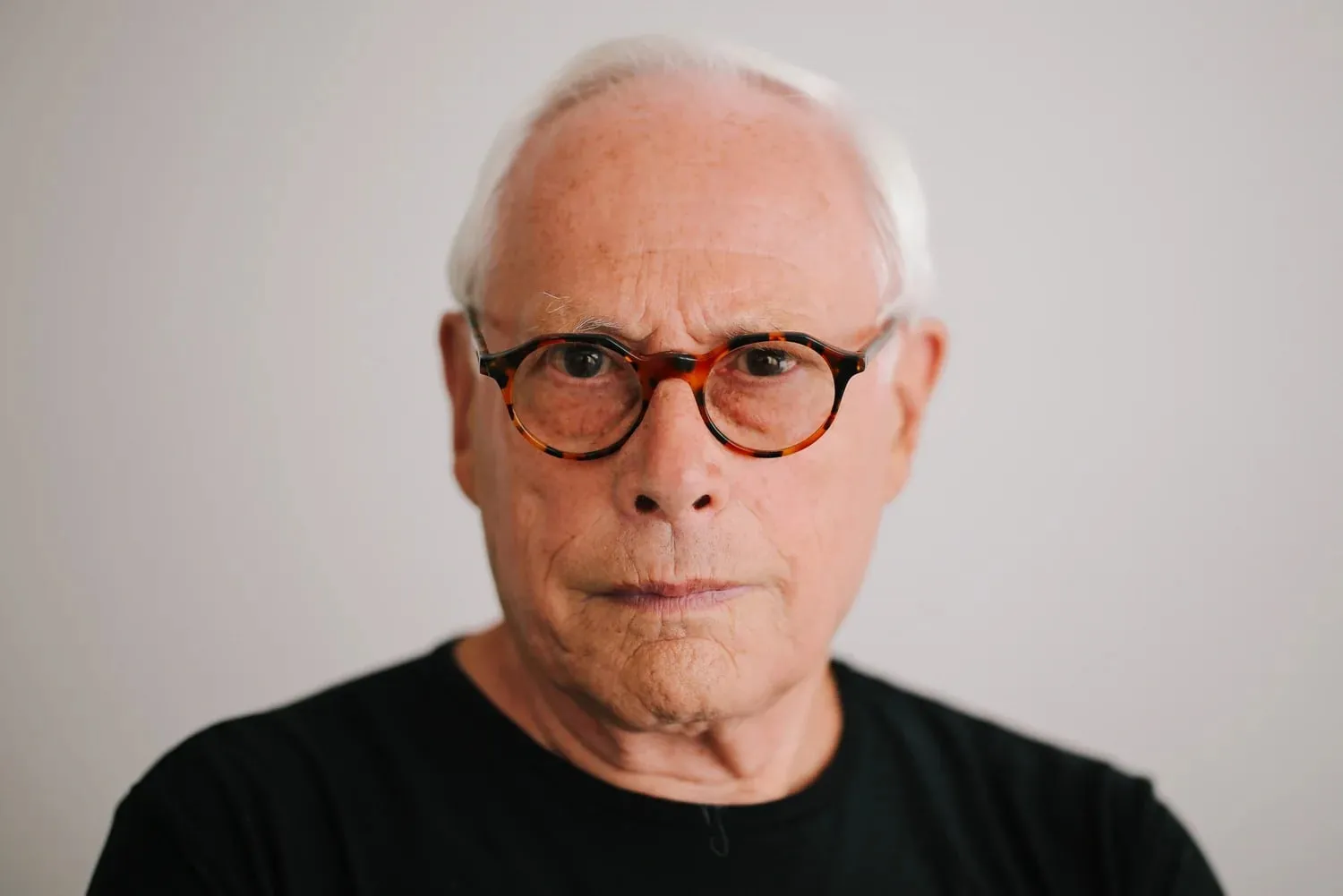 Retrato de Dieter Rams con gafas de pasta y camiseta negra sobre fondo gris