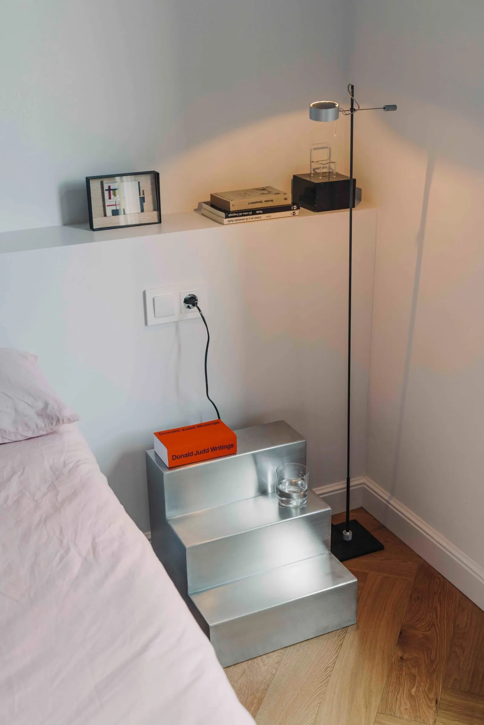 Mesa auxiliar Escala de acero junto a una cama con rosa pálido, con un libro naranja y un vaso de agua en sus peldaños y una lámpara de pie al lado