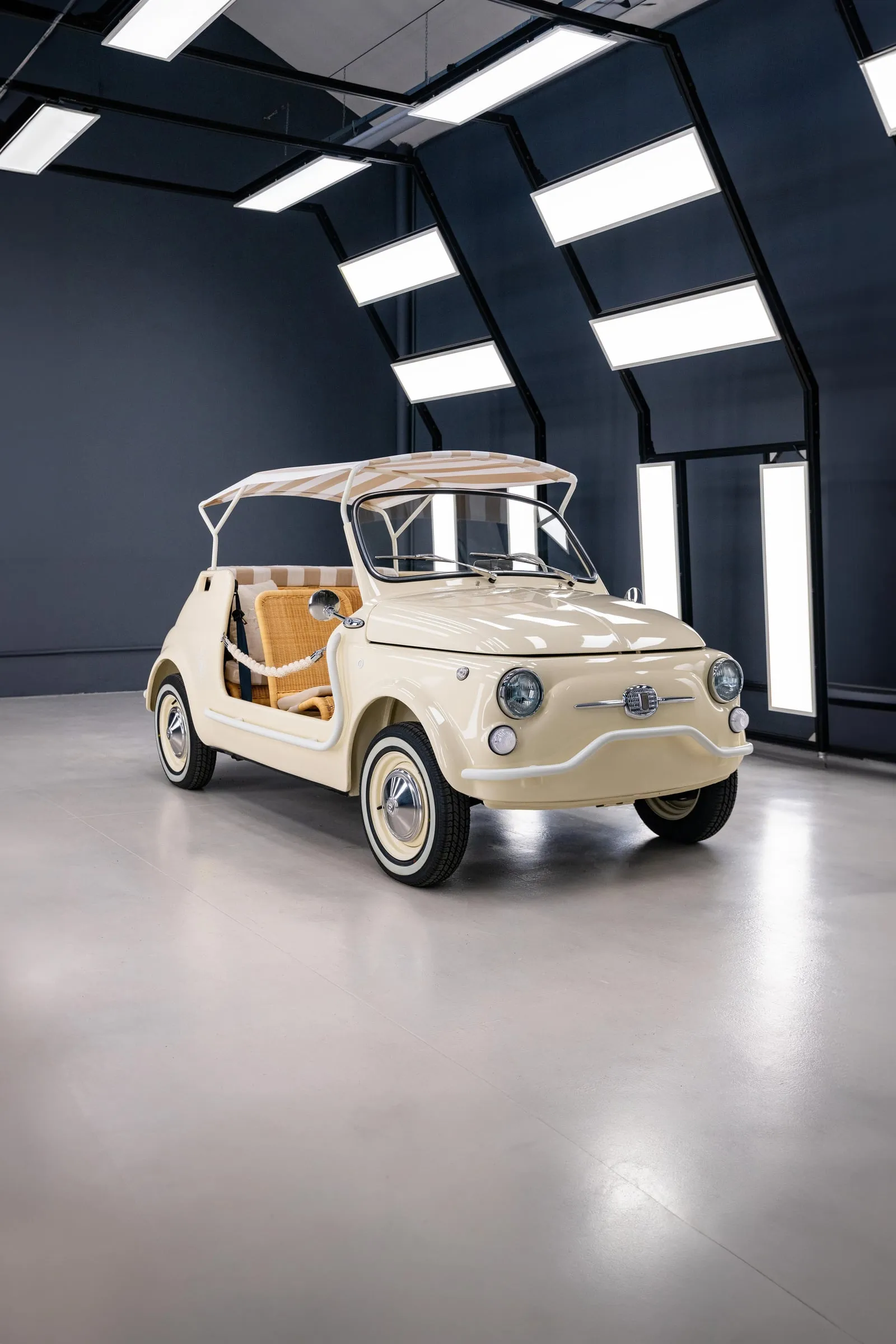 Fiat 500 Spiaggina color crema en una nave oscura de paneles luminosos, visto de frente en tres cuartos, con capota de rayas sobre una estructura fina