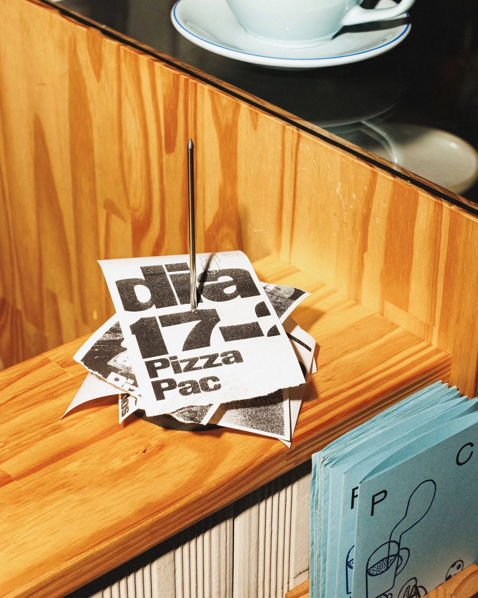 Montón de papeles pequeños con el texto «dia 17, Pizza Pac» sobre un mostrador de madera, junto a bolsas azul claro