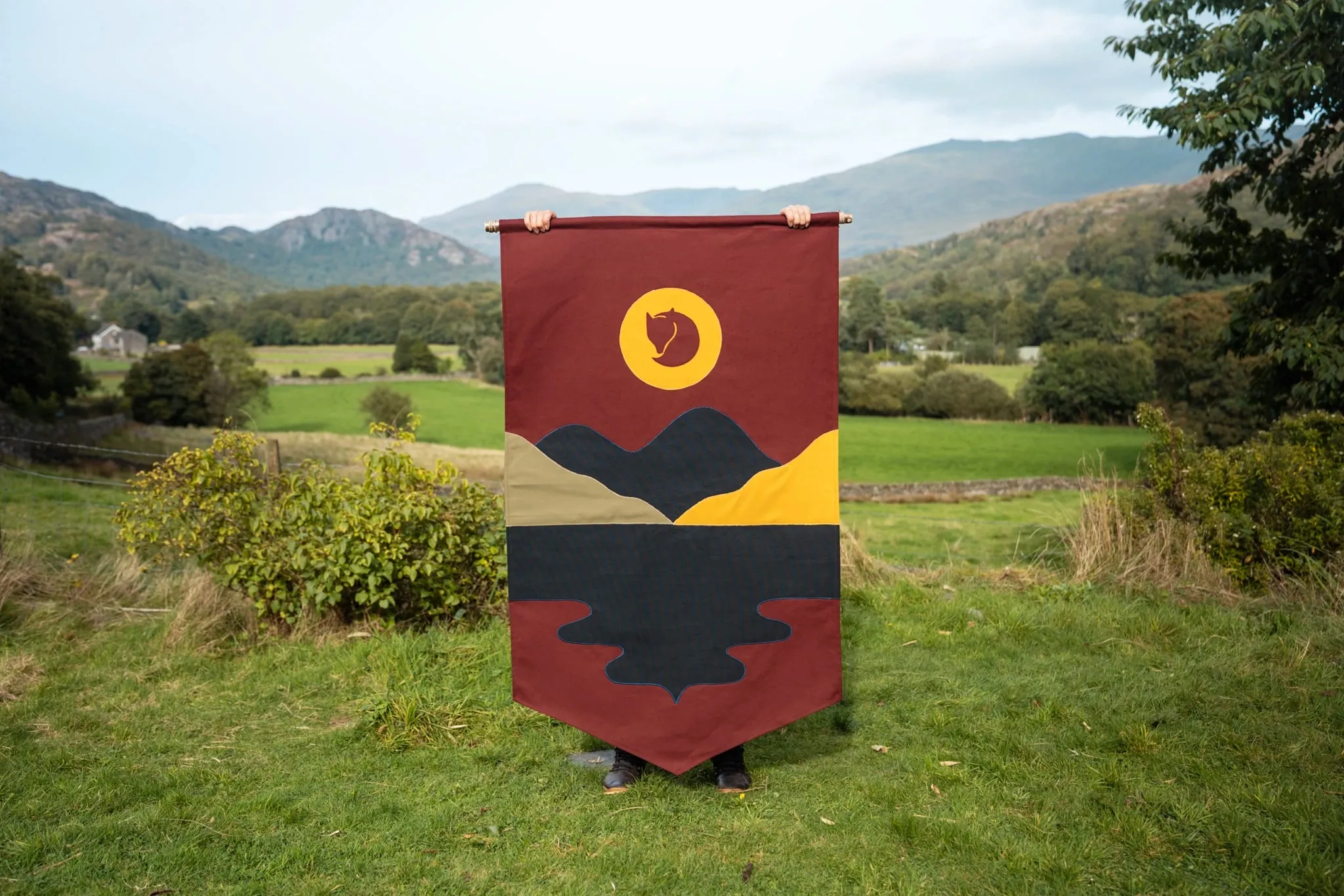 Bandera granate con un sol amarillo, montañas y un lago negro con su reflejo cosidos con tela
