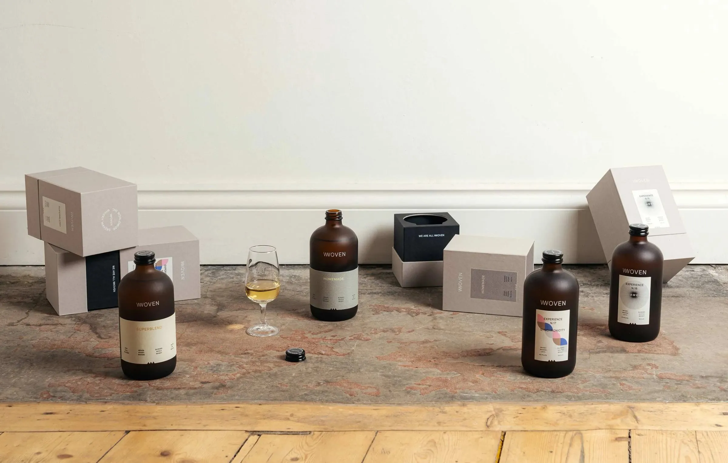 Seis cajas de cartón gris, botellas de Woven y una copa con whisky sobre el suelo de una sala