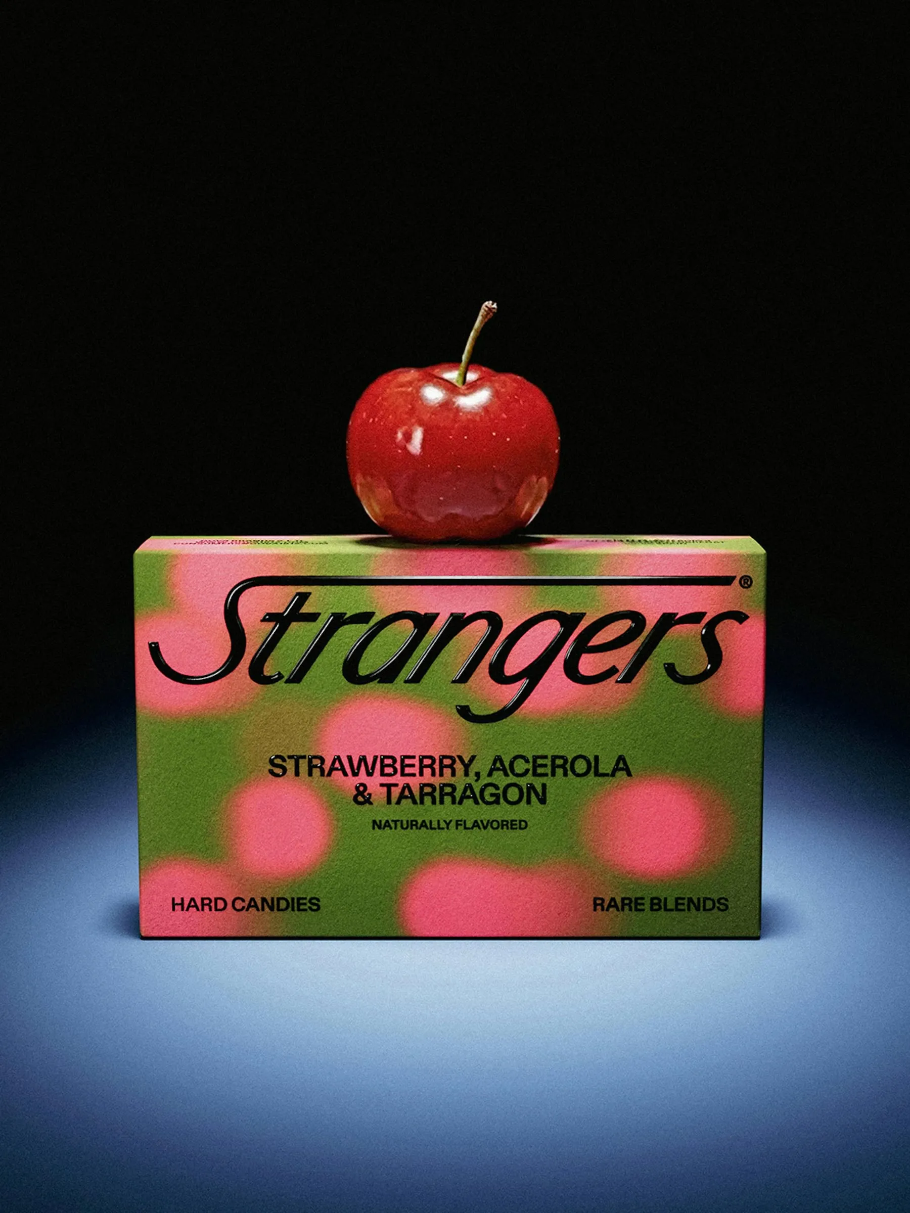 Caja verde de Strangers con manchas rosas, de fresa, acerola y estragón, con una manzana roja encima sobre fondo negro