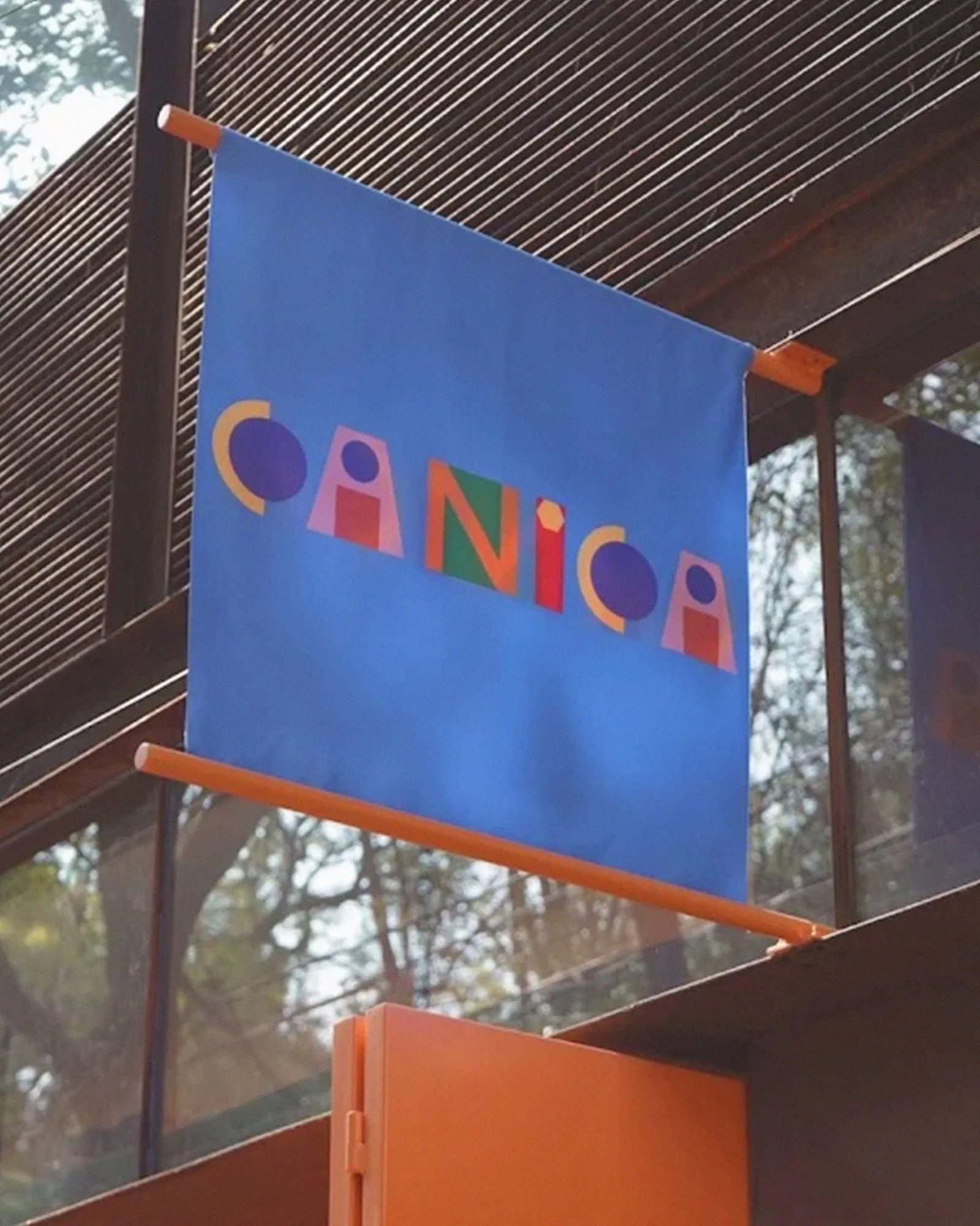 Rótulo de tela azul con el logotipo de Canica colgado de dos barras naranjas en la fachada del café