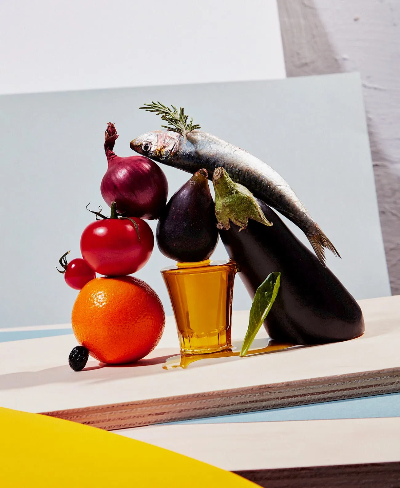 Bodegón de una sardina en equilibrio sobre una cebolla roja, un higo y una berenjena, junto a tomates, una naranja y un vaso de aceite de oliva derramado