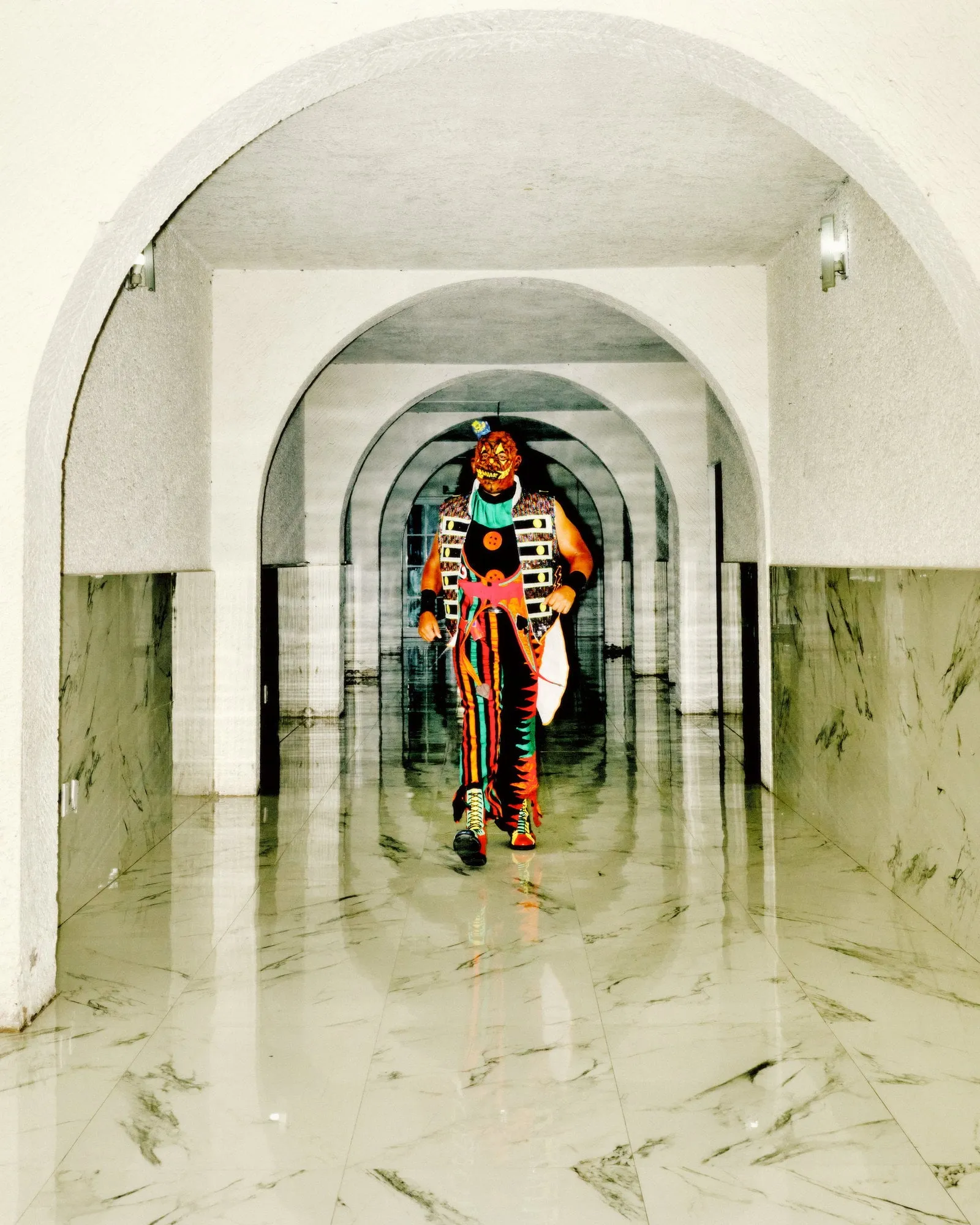Luchador con máscara y traje de colores camina solo por un pasillo de arcos y suelo de mármol reflectante