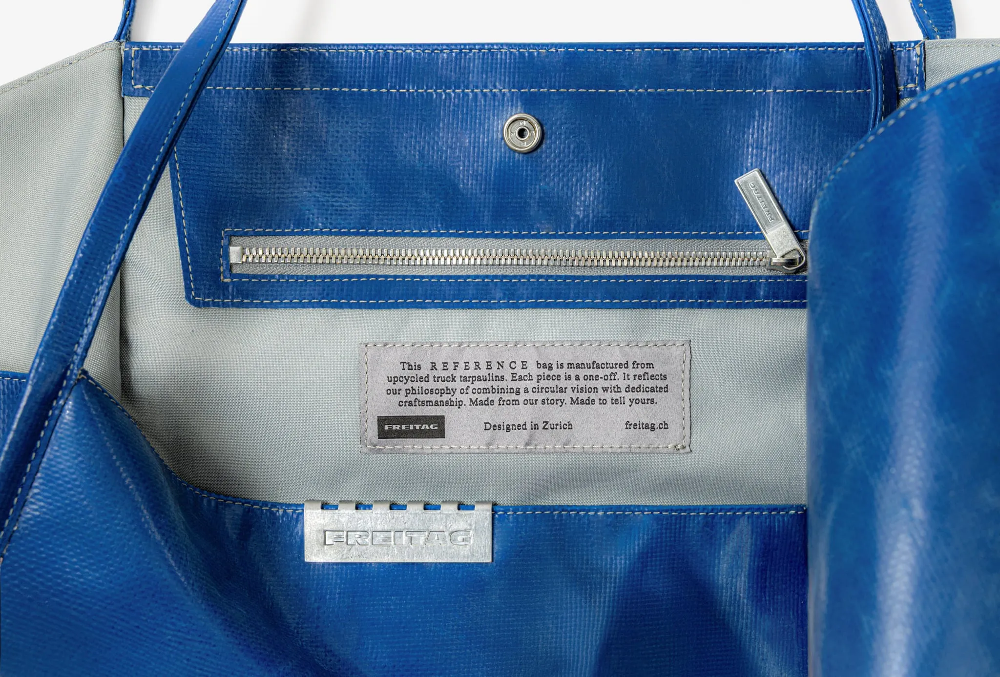 Interior de un tote azul con bolsillo con cremallera, la etiqueta de tela gris que explica el material y la chapa metálica de Freitag
