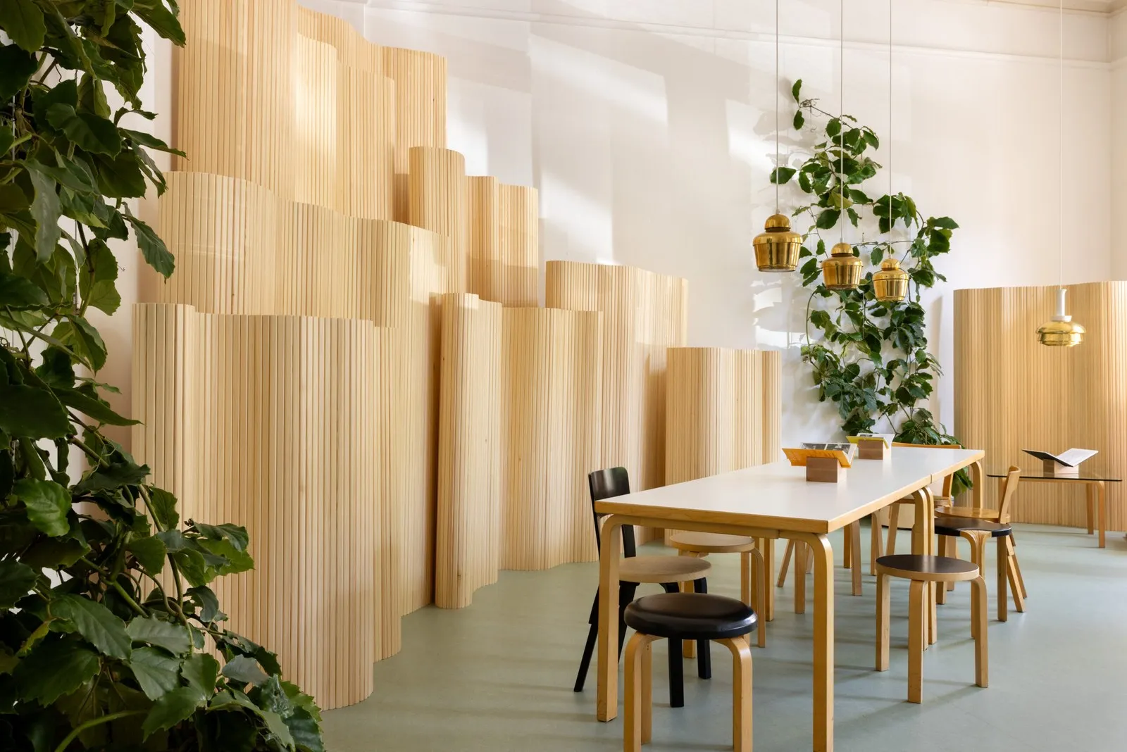 Sala con paneles de madera clara ondulados, una mesa blanca con taburetes y tres lámparas doradas colgadas