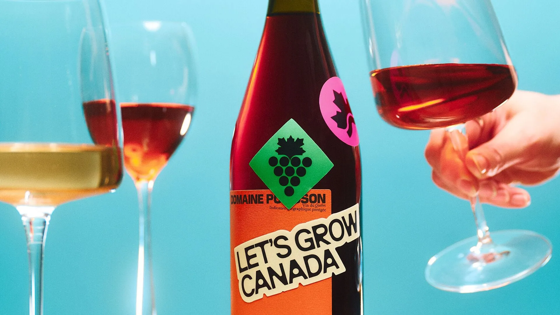 Una botella de vino tinto con una pegatina verde en forma de rombo con un racimo de uvas rematado por una hoja de arce, y otra pegatina rosa con el logotipo