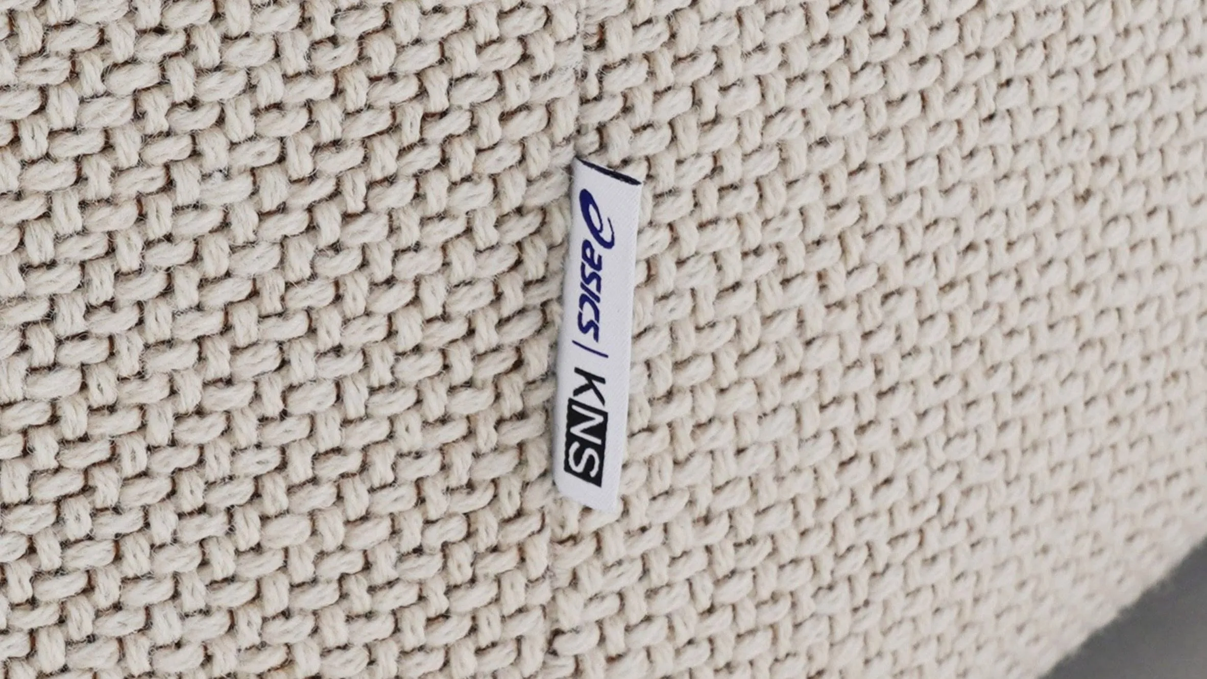 Primer plano de una tela de trama gruesa color crema con una etiqueta blanca bordada con los logotipos de ASICS y KNS