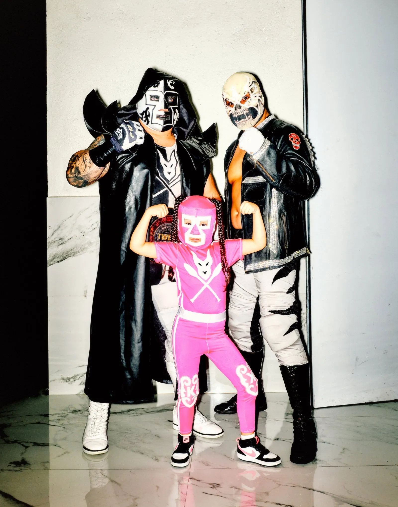 Tres luchadores enmascarados, dos adultos y una niña con mono rosa, posan flexionando los brazos en un interior de mármol