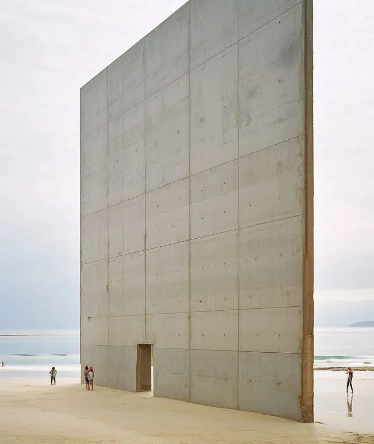 Muro gigante de hormigón con una pequeña puerta, plantado sobre la arena de una playa, con varias personas diminutas paseando a su alrededor