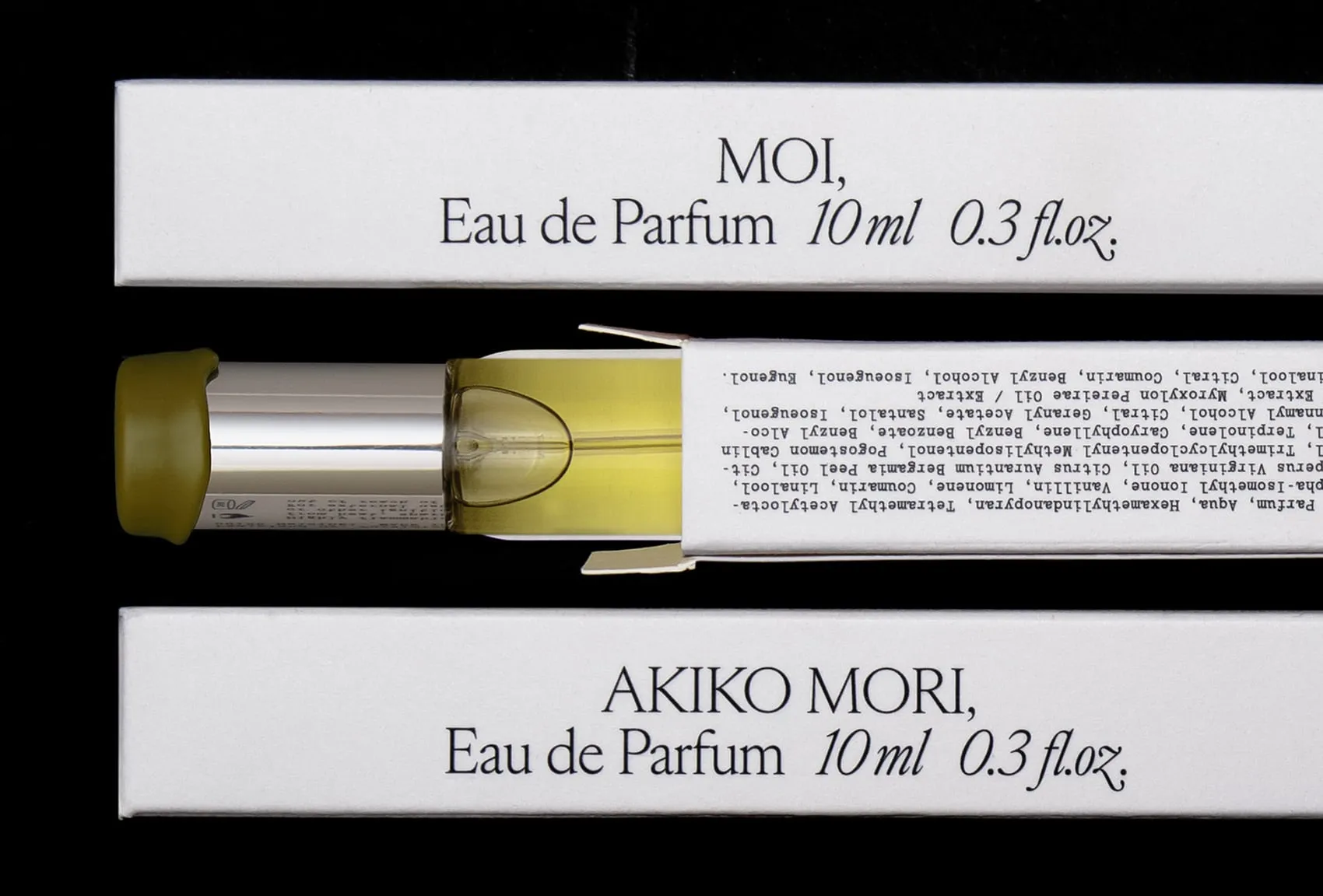 Frasco con tapa de cera verde oliva y cajas de 10 ml de Moi y Akiko Mori sobre fondo negro, con la lista de ingredientes impresa a máquina