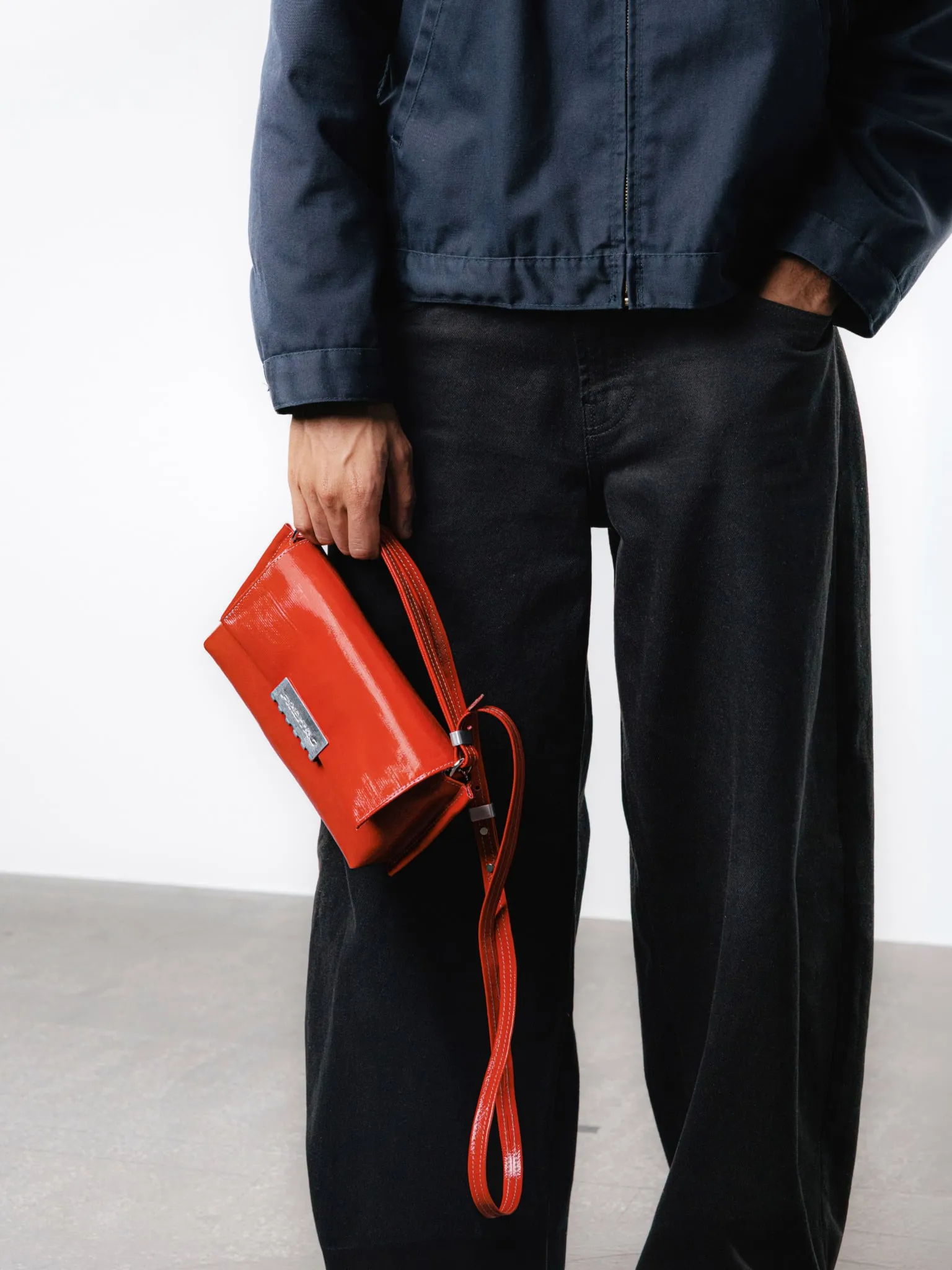 Mano con un bolso cruzado rojo brillante colgado de una correa larga, ante una persona vestida de azul marino y vaqueros negros