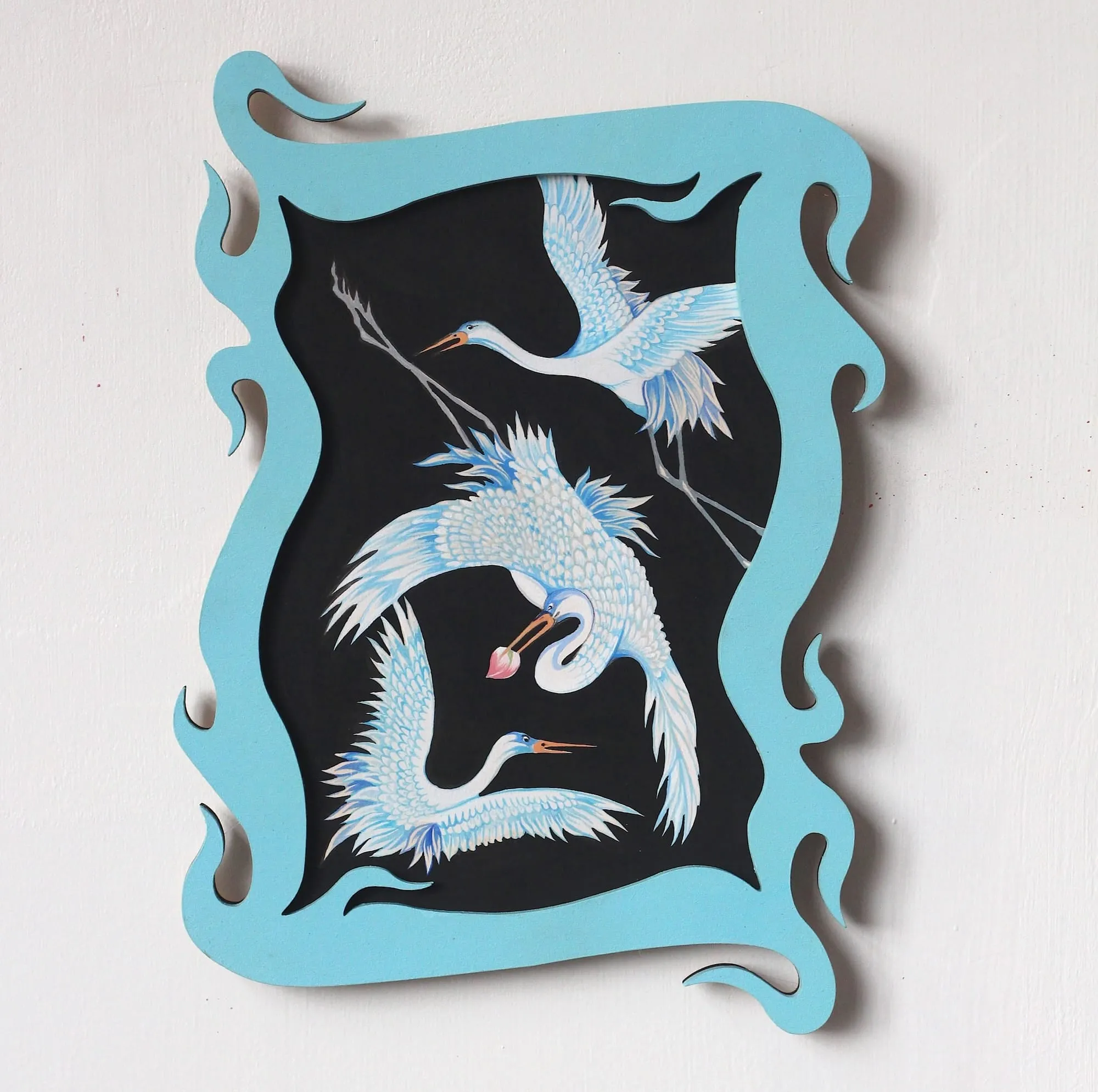Panel en forma de llama azul claro con tres grullas blancas, una con una flor de loto en el pico, sobre fondo negro