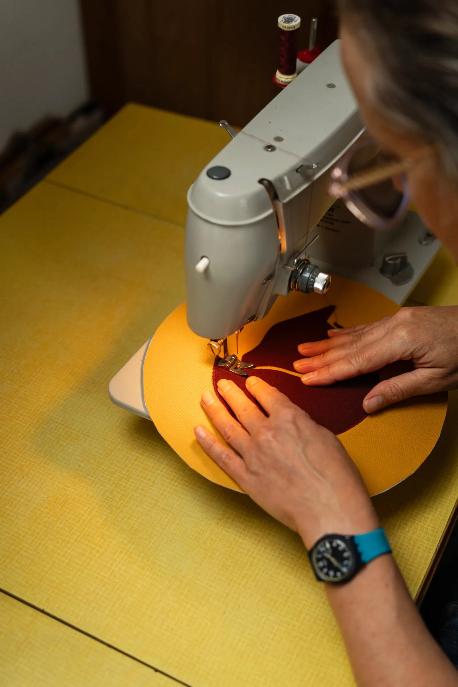 Manos que guían una tela amarilla bajo la aguja de una máquina de coser, con hilo granate